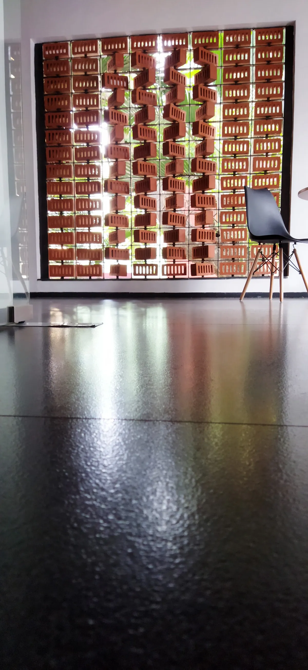 A terracotta brick jaali screen filters daylight and greenery into an entrance area with a single black shell chair and reflective dark flooring.