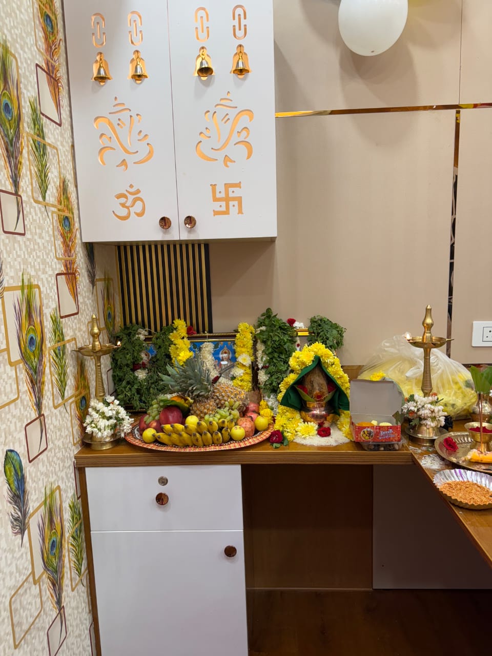 Pooja unit with a laser-cut white cabinet featuring Om and Ganesha motifs, peacock-feather wallpaper on the side wall