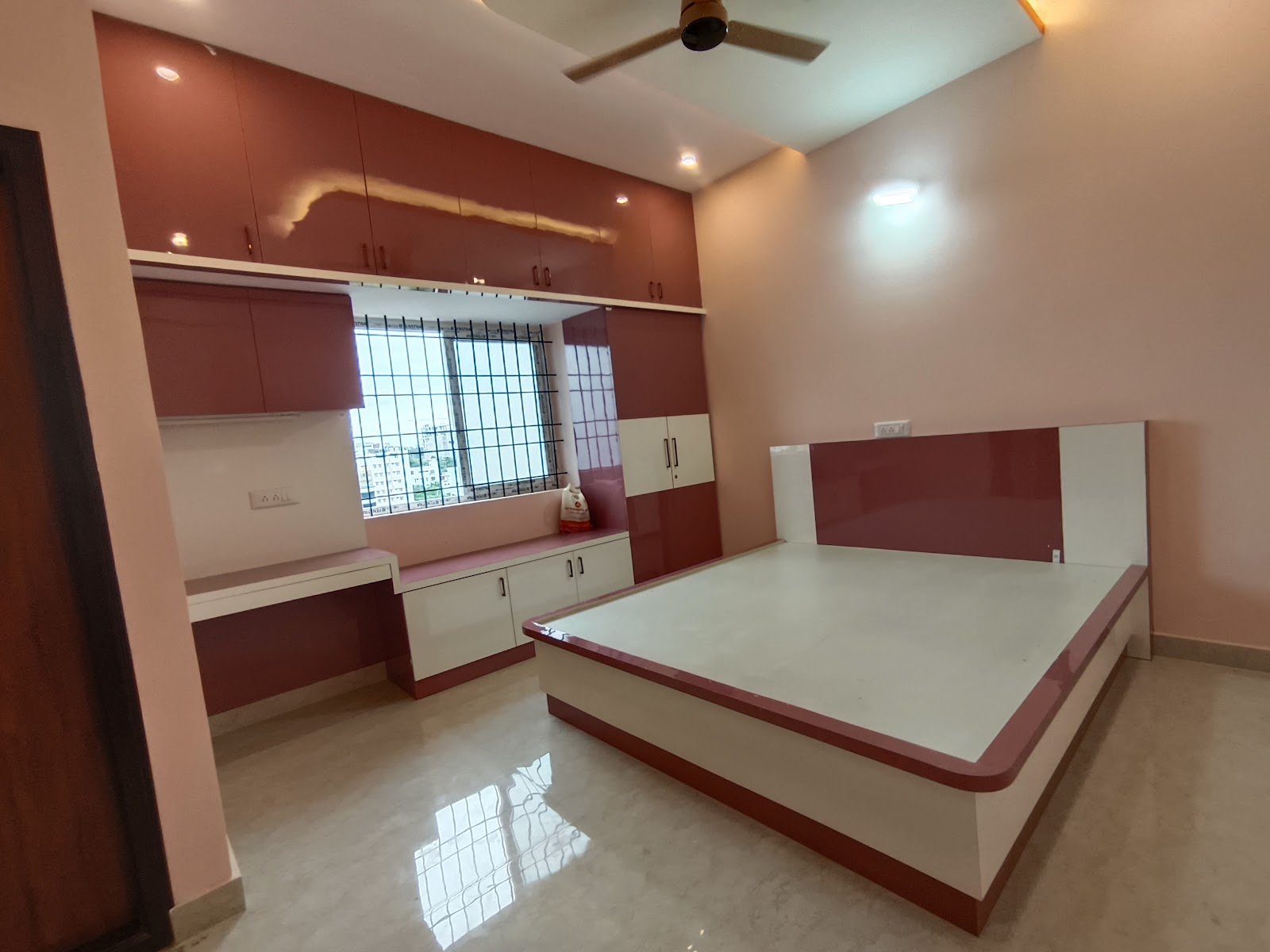 Under-construction bedroom with a glossy maroon-and-white built-in bed platform, matching wardrobe, and a window-side study counter.