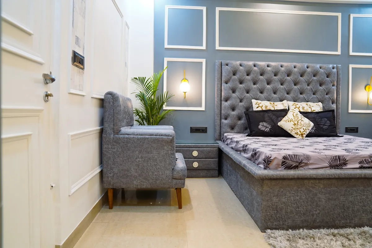 Grey velvet button-tufted bed against a slate-blue wall gridded with white moulding frames, brass-and-glass sconces inside two of the frames
