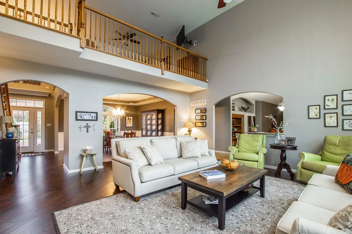Real photo of a double-height living room with a wood-balustraded loft above, arched openings to a dining room and kitchen, cream sofas