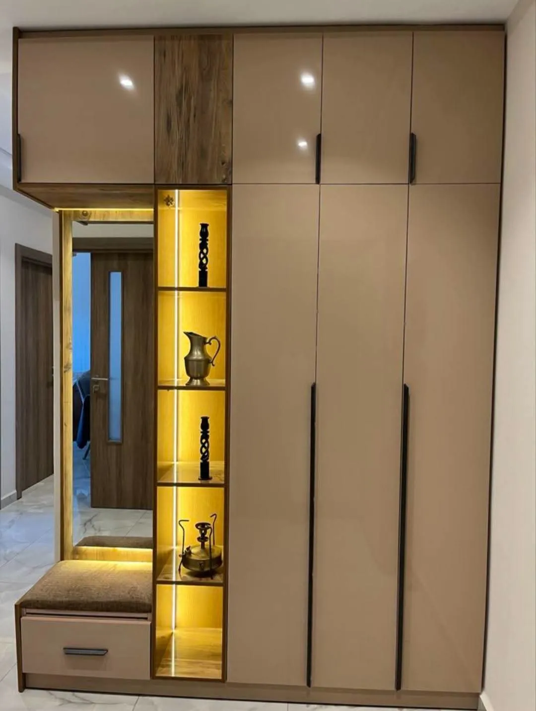 Entrance foyer wall combining a full-height taupe wardrobe with a backlit brass-vase display niche, a mirror, and an upholstered bench with a drawer.