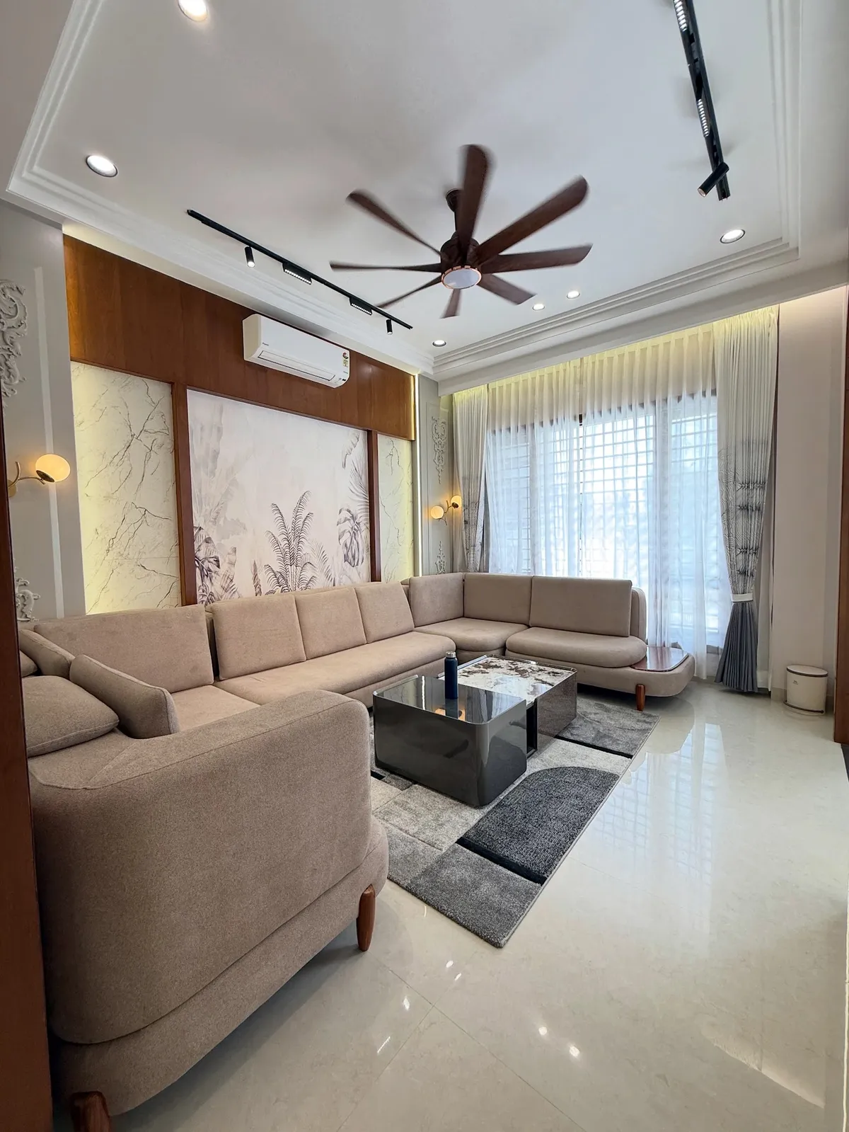 Beige U-shaped sectional and a smoked-mirror coffee table on a grey block rug, facing a backlit tropical-leaf mural framed in teak