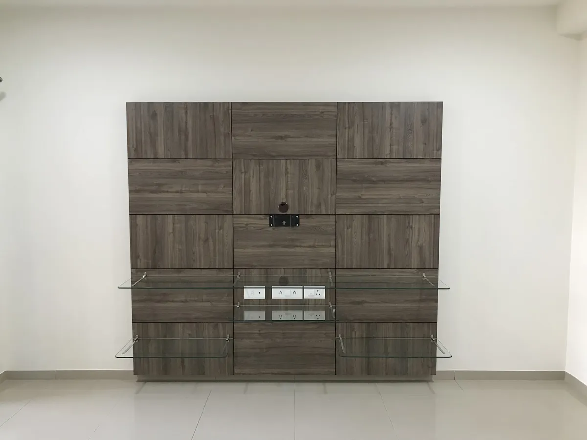 A walnut-toned wall-panel TV unit with glass display shelves and concealed wiring points, on an otherwise bare living-room wall.