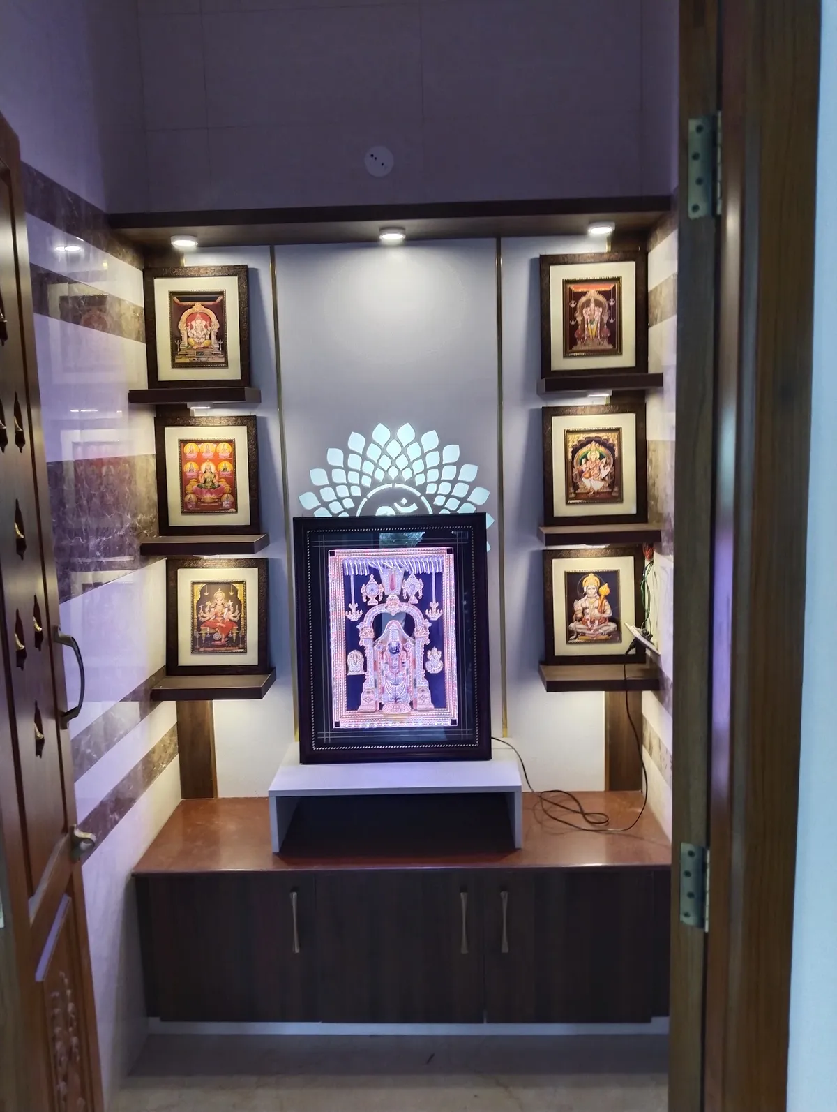 Niche pooja unit behind carved teak doors, with six framed deity pictures stepped on wenge floating shelves either side of a backlit acrylic Om-and-lotus motif