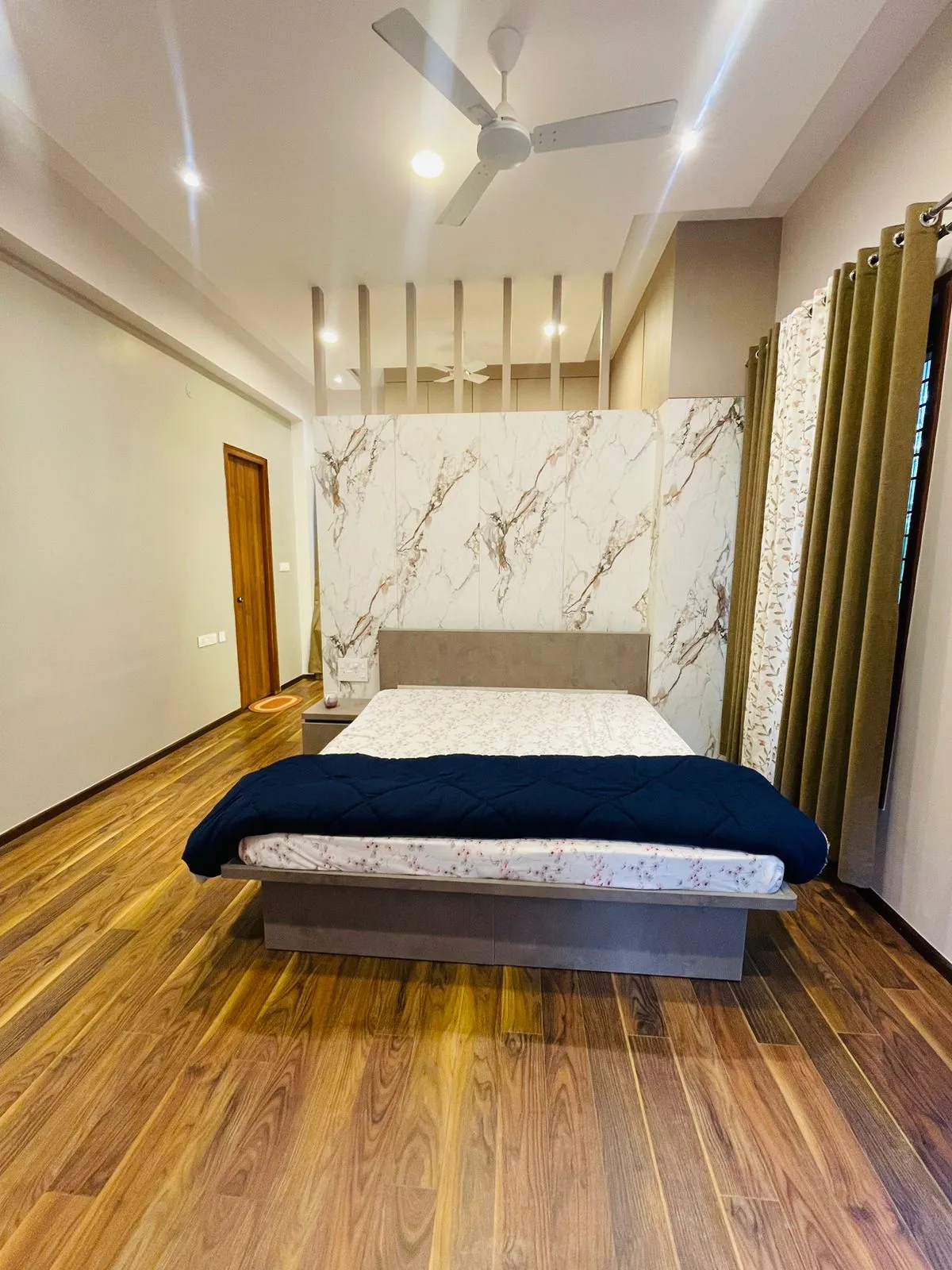 Bedroom with a book-matched white marble-look slab wall behind a low concrete-finish platform bed, olive eyelet curtains over floral sheers