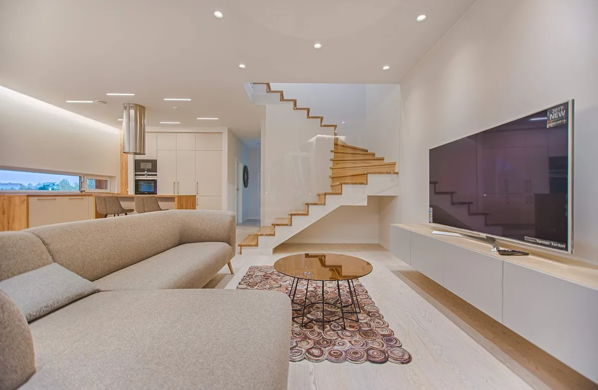 Pale oak duplex living room where a floating white console runs the full length of the TV wall and a cantilevered timber-tread stair with a glass balustrade