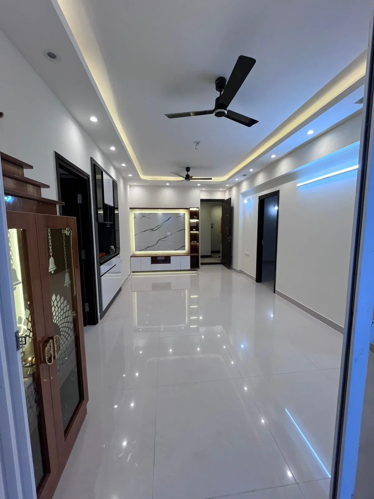 Handover photo of an unfurnished flat living hall: a white-and-walnut TV unit under a backlit veined marble panel, mirror-gloss white floor tiles