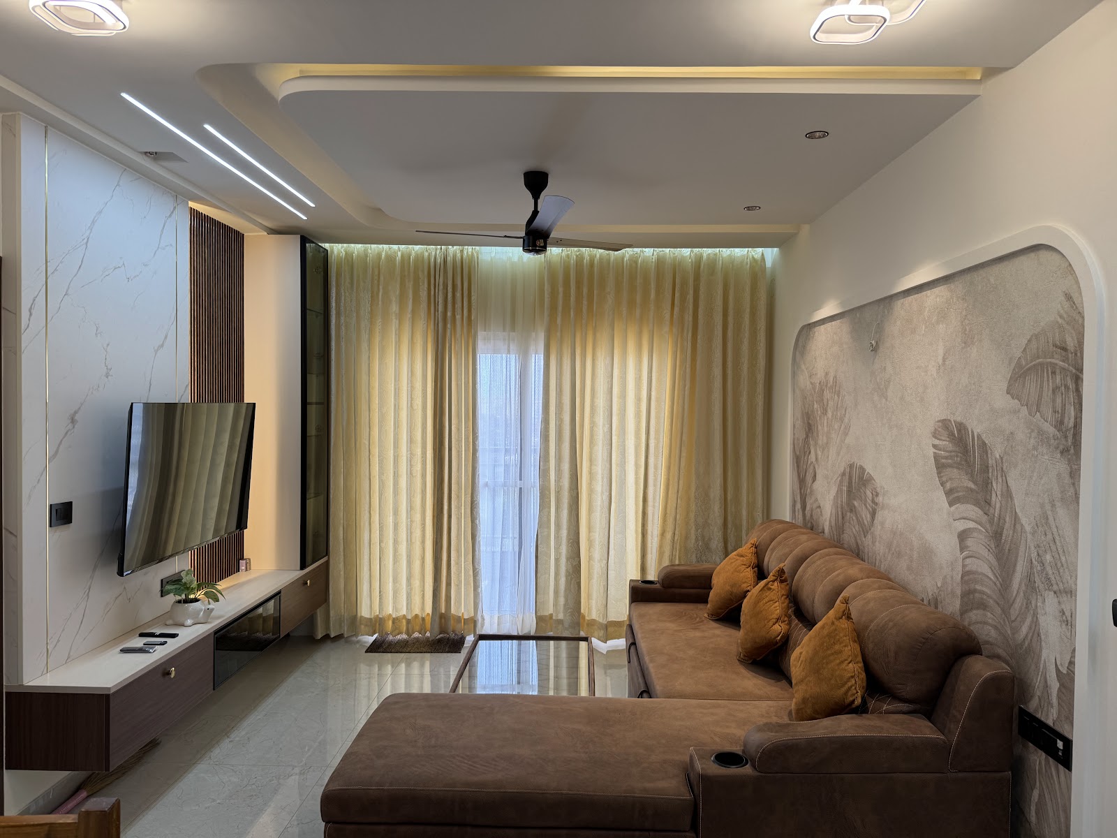 Real photo of a living room with a marble-clad TV wall, a botanical mural wallpaper behind a brown leather reclining sectional