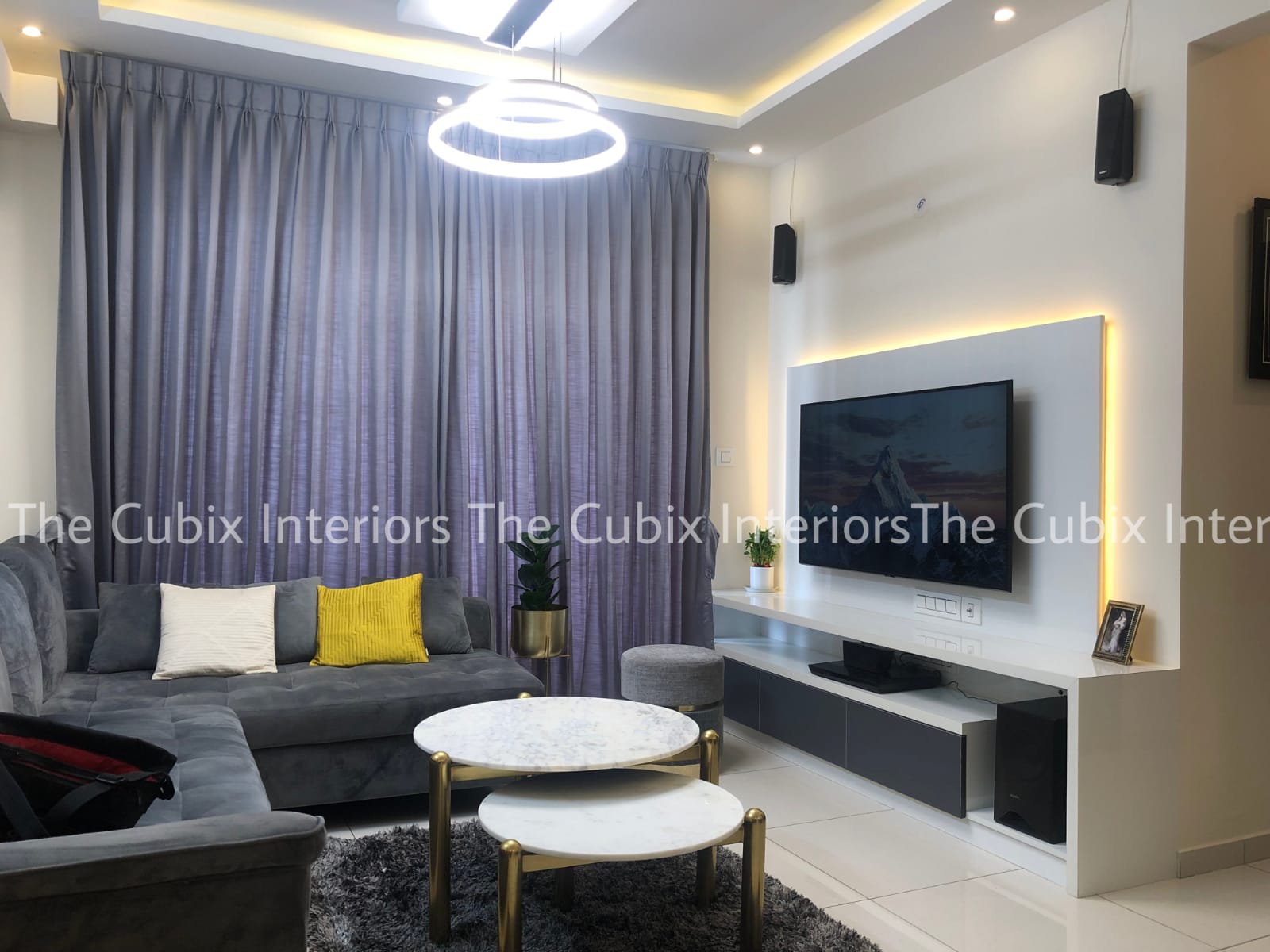 Living room with a grey L-shaped sectional sofa, yellow and white cushions, a backlit white TV feature wall, and round marble-and-gold nesting coffee tables.
