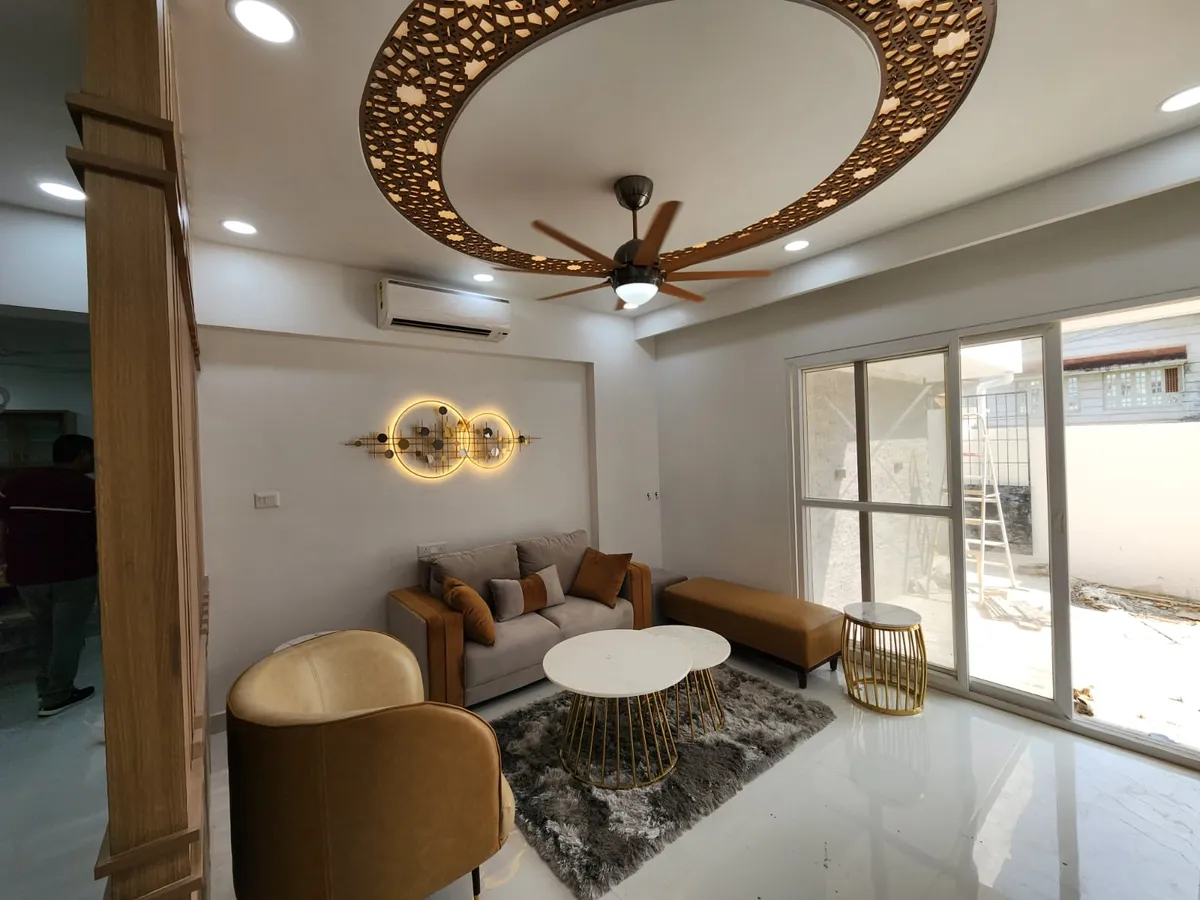 Living room under a large circular ceiling drop ringed with backlit CNC-cut walnut jaali, with a wooden-bladed fan at its centre over a beige sofa