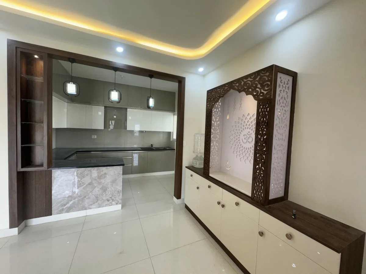 Backlit carved-wood jaali pooja unit with an Om motif beside a foyer opening onto a glossy modular kitchen with pendant lighting.