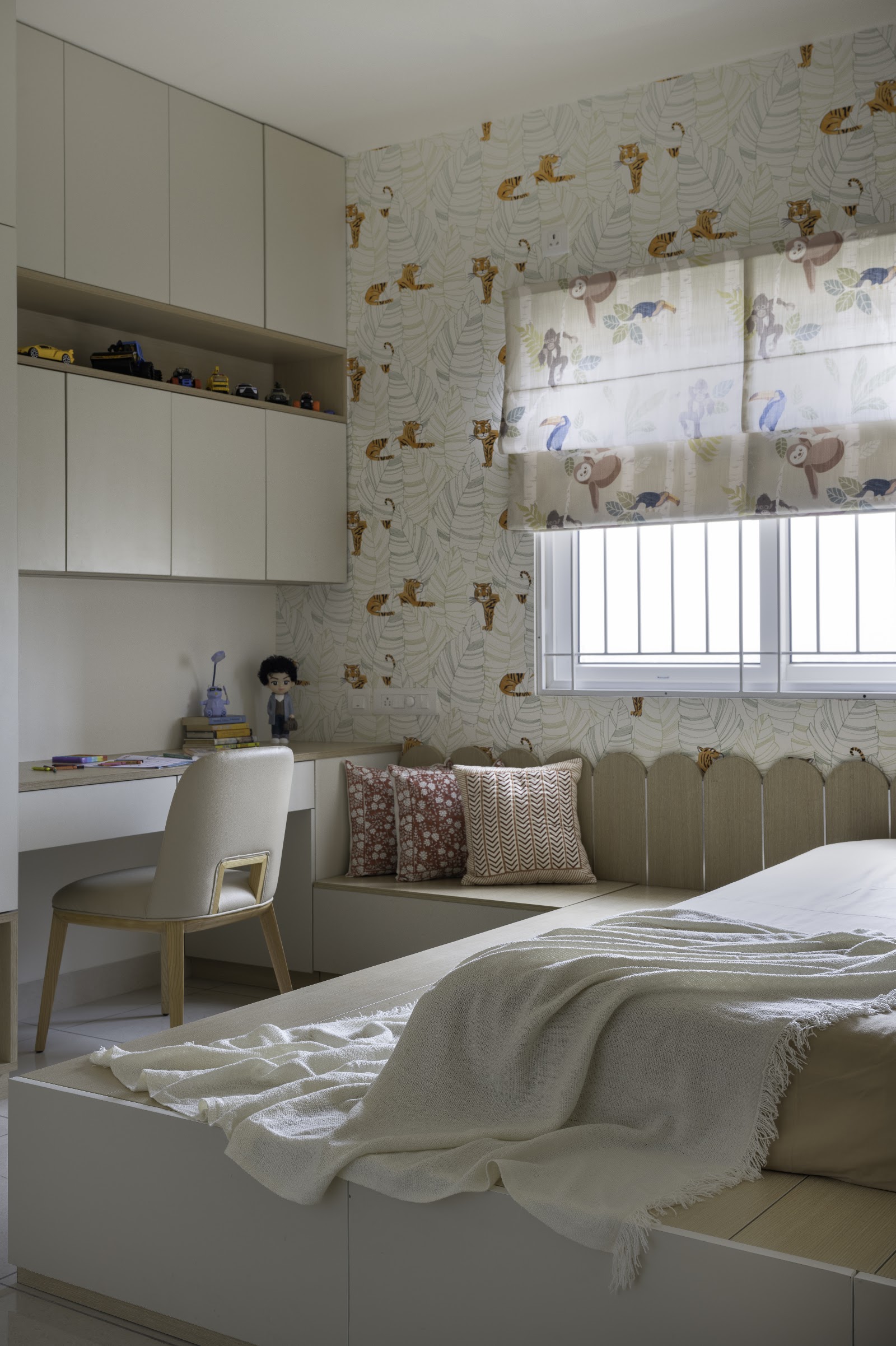 Kids' bedroom with jungle-animal wallpaper and a matching roman blind, a built-in window seat with cushions, and a study nook styled with toy cars.