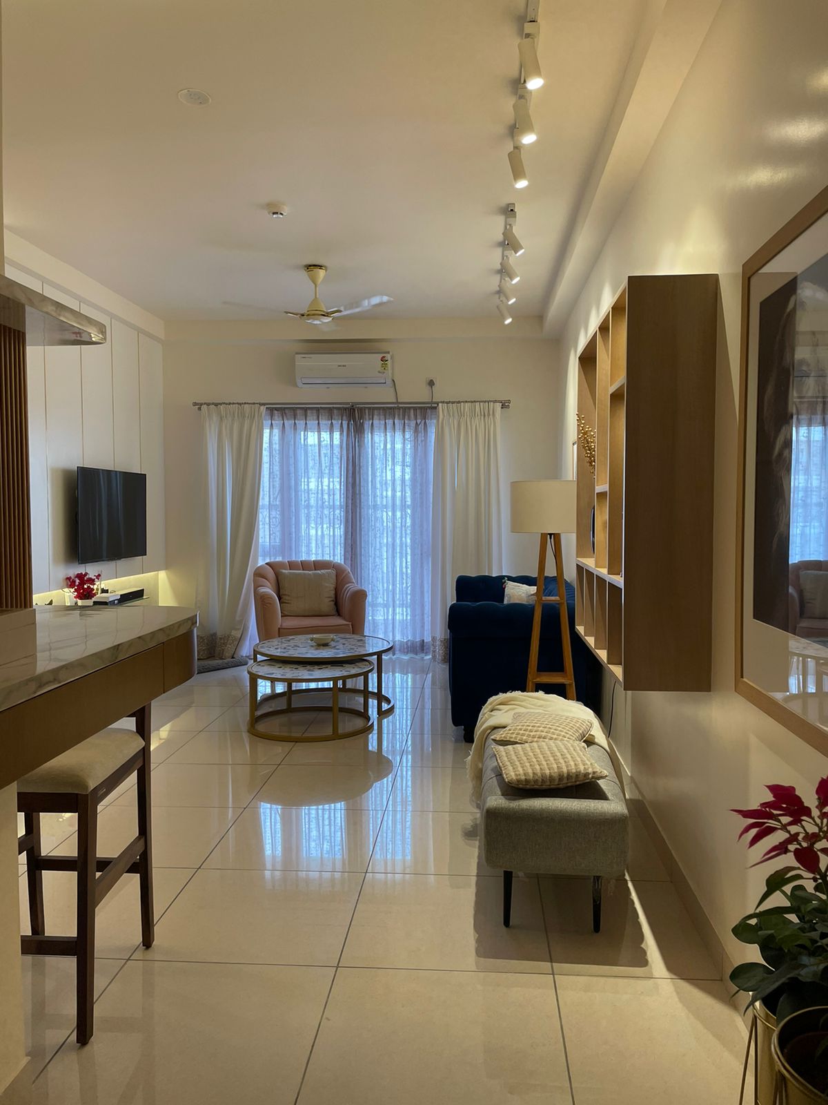 Living room with a navy velvet sofa, a blush pink accent armchair, nested marble-top coffee tables, and an open wood shelving unit beside sheer curtains.