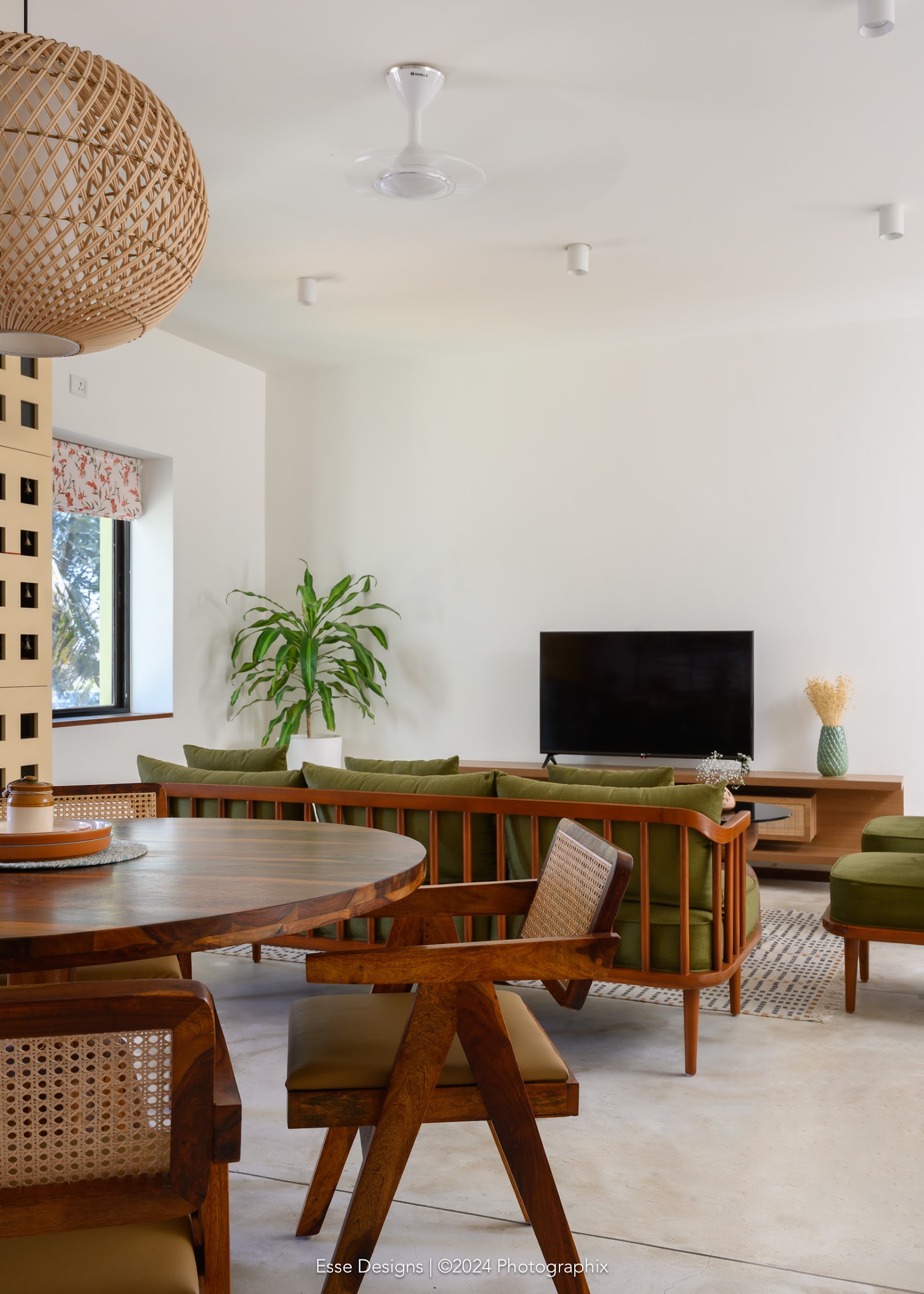Open-plan dining and living area with a round wood table and cane-back chairs in the foreground, a green bench-style sofa and wood TV console beyond