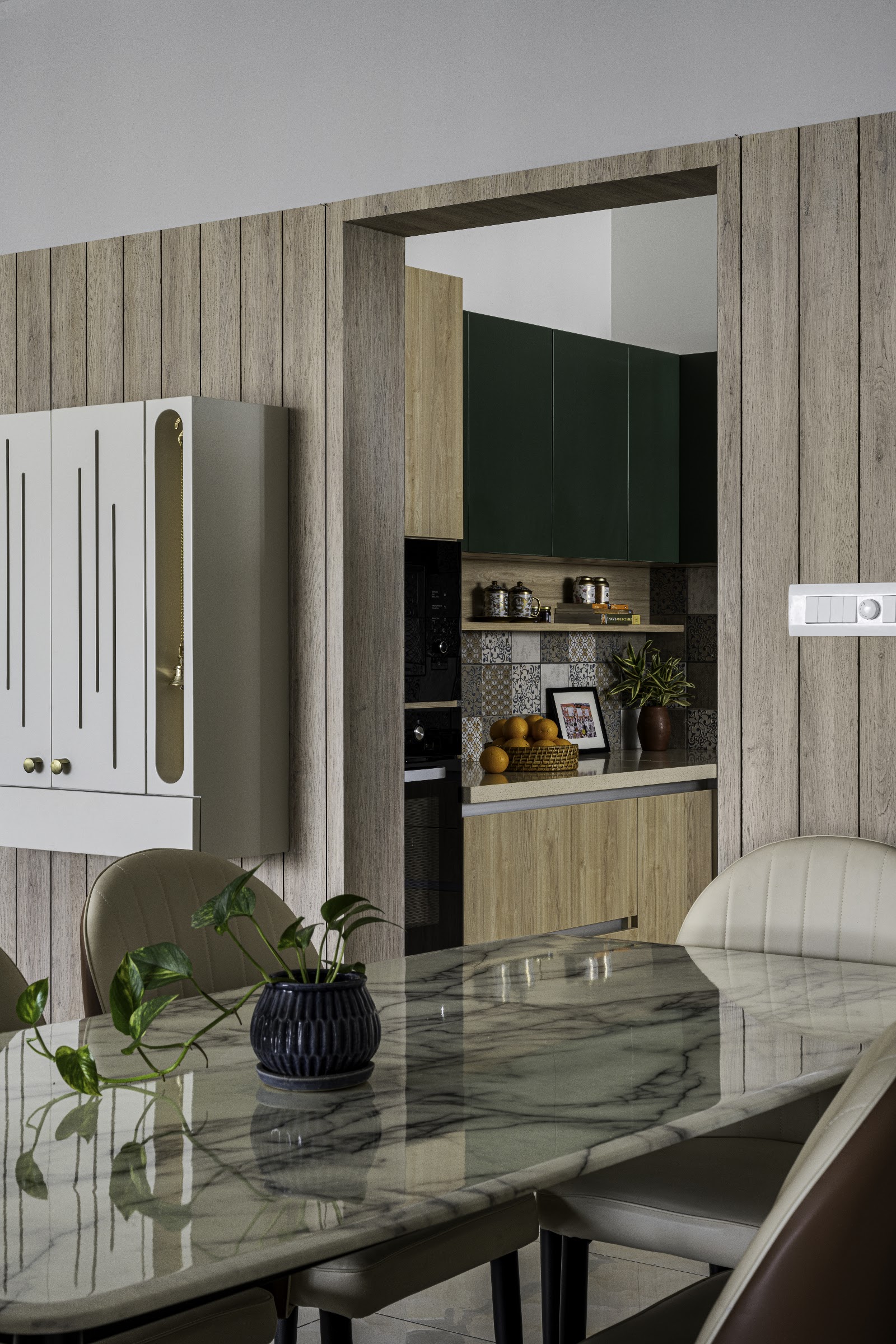Marble-top dining table with tan leather channel-back chairs in the foreground, opening through a wood-panelled doorway to a dark green modular kitchen