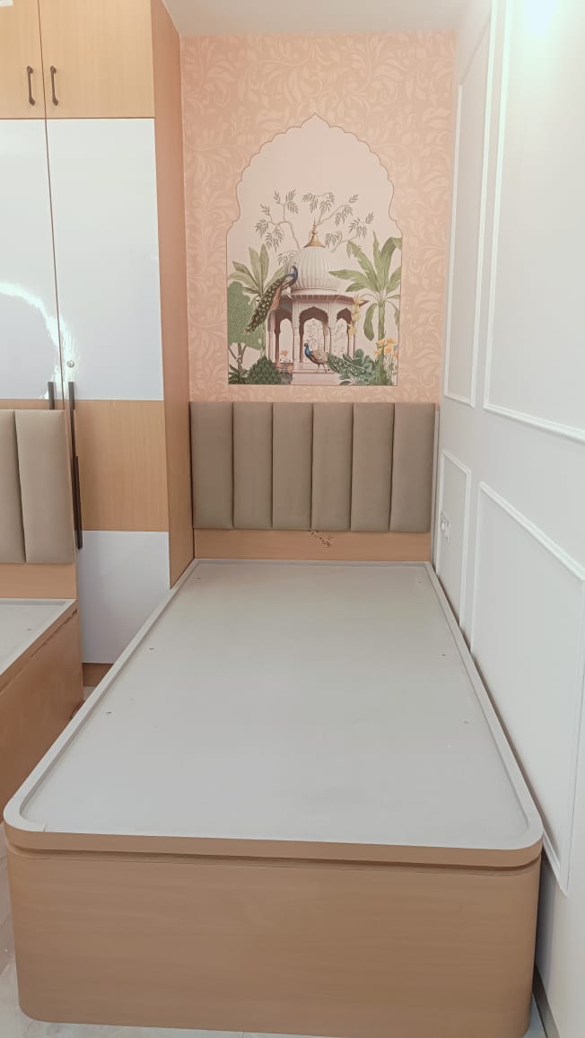 Single storage bed with a fluted taupe upholstered headboard against a pink wallpaper wall featuring a peacock and palace mural, beside a laminate wardrobe.
