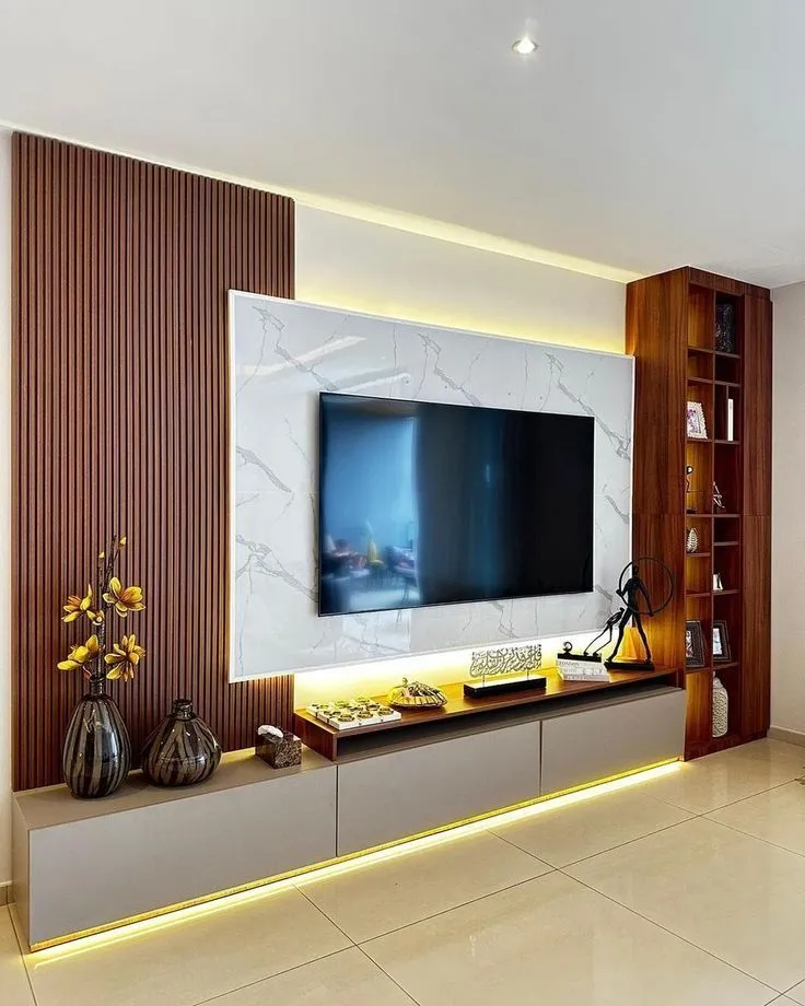 Living-room TV wall where a white-veined marble-look panel carrying the TV sits between a full-height fluted teak wall on the left and a slim teak open-shelf