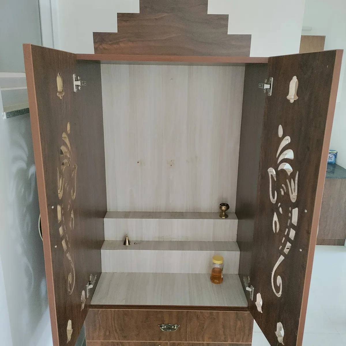 Open wooden pooja unit cabinet with carved brass-inlay door panels and a two-tier idol shelf, installed beside a kitchen counter