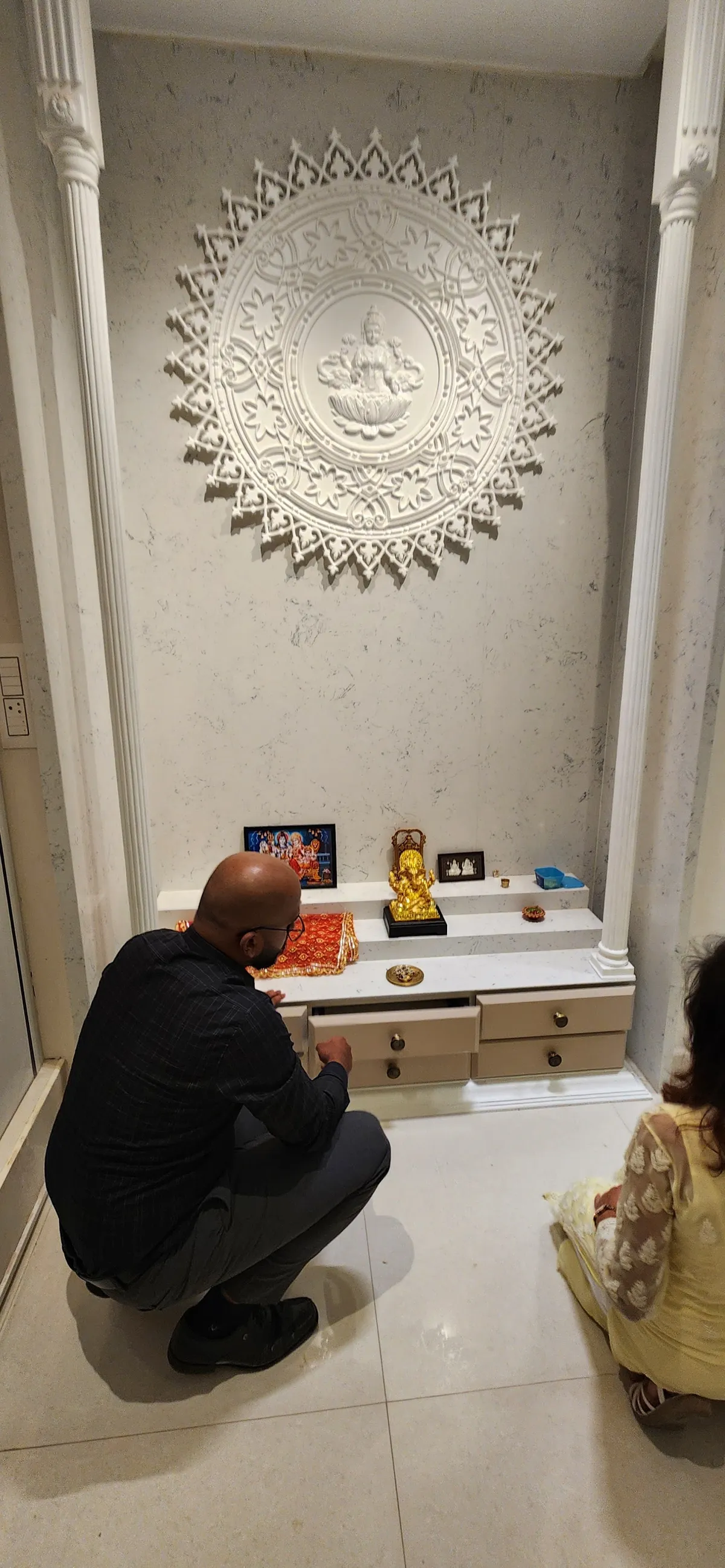 Marble-clad pooja niche with a carved white Lakshmi medallion between fluted pillars, a family at prayer before the drawer-fronted counter.