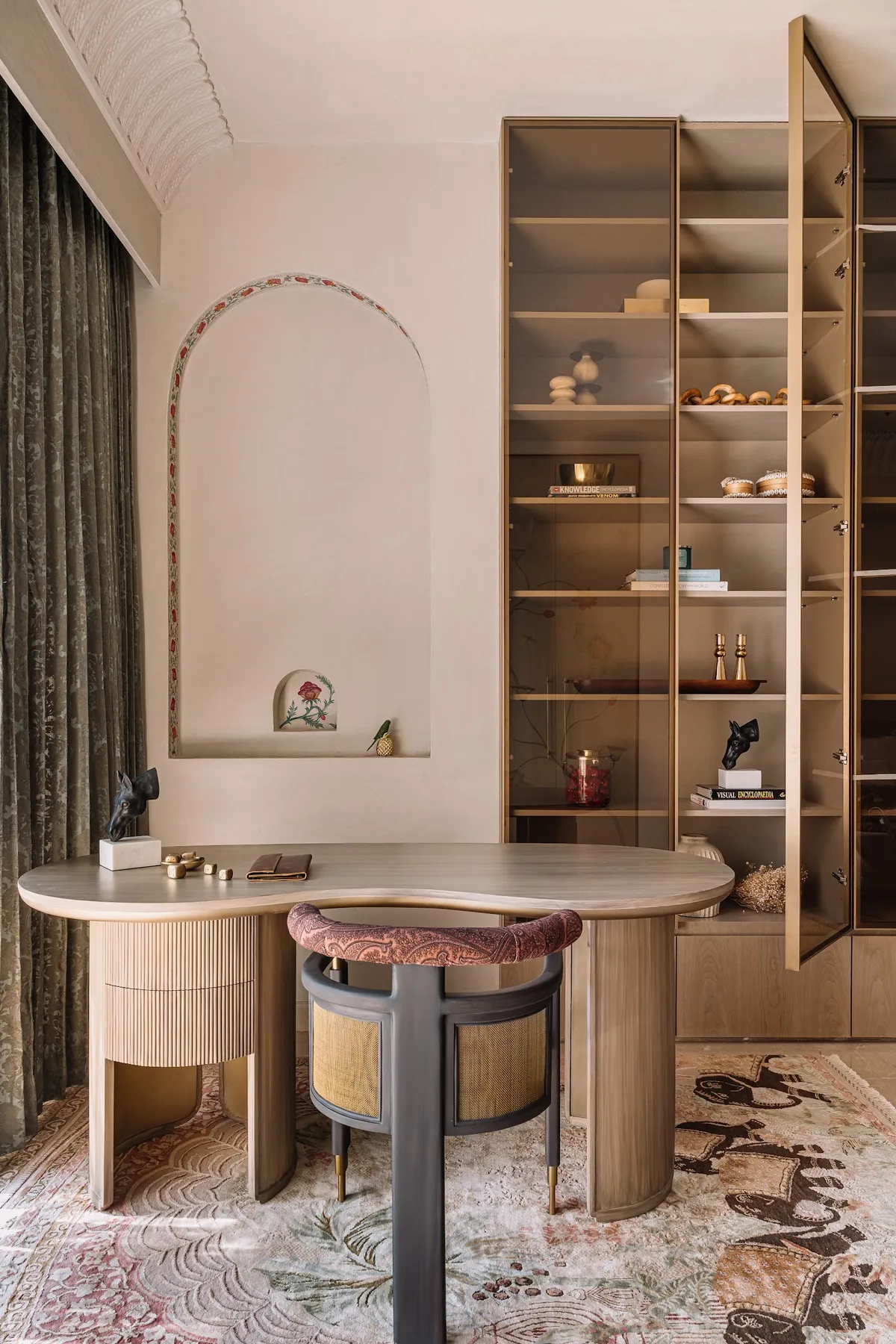 Study nook with a sculptural kidney-shaped wood desk, a cane-back accent chair, and a brass-framed glass display cabinet beside a hand-painted arched wall