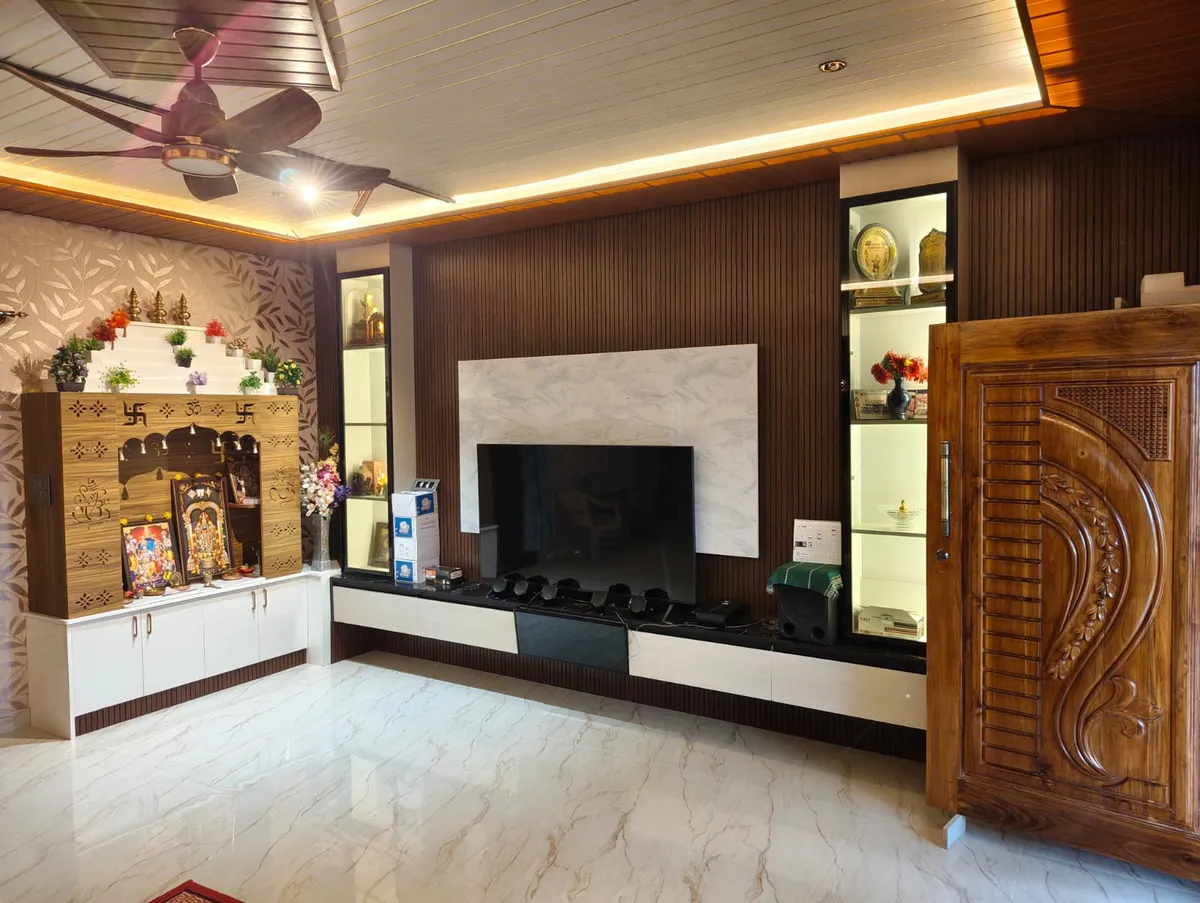 Living room where a dark fluted TV wall with a white marble slab and two backlit glass display columns meets a carved teak pooja mandir stepped in white