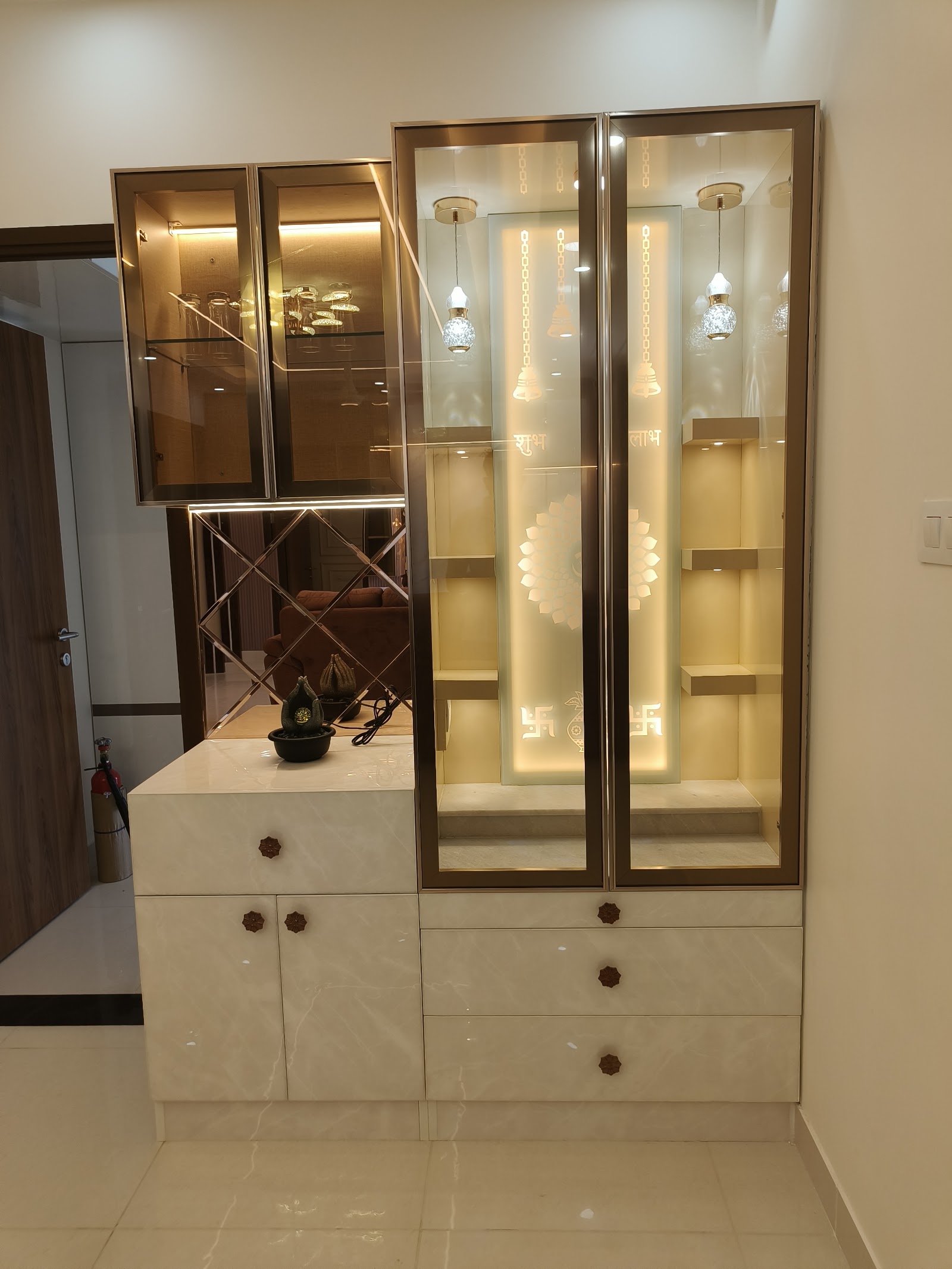 Backlit glass pooja unit with etched 'Shubh Labh' script and hanging bell motifs, paired with a mirrored crockery display cabinet and marble-top console below.