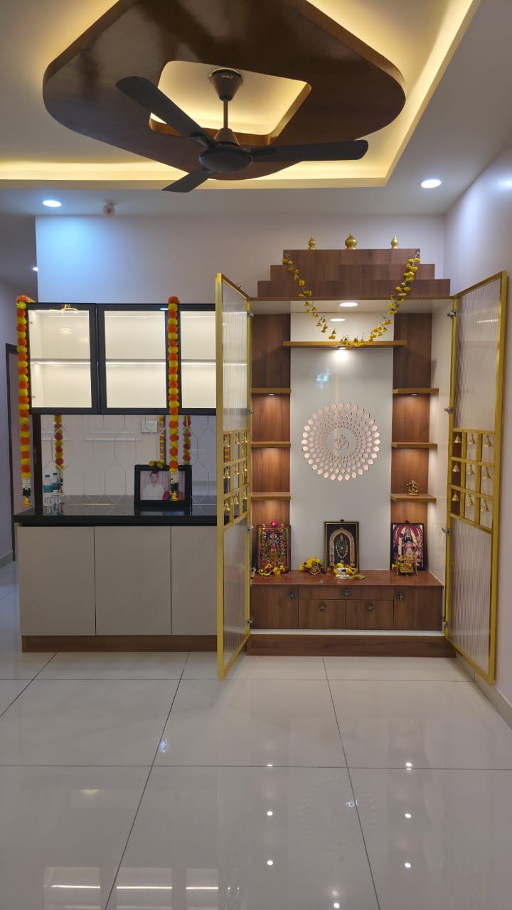 Dedicated pooja room with a tiered wooden mandir unit, a backlit Om medallion niche, gold-trimmed glass doors hung with bells