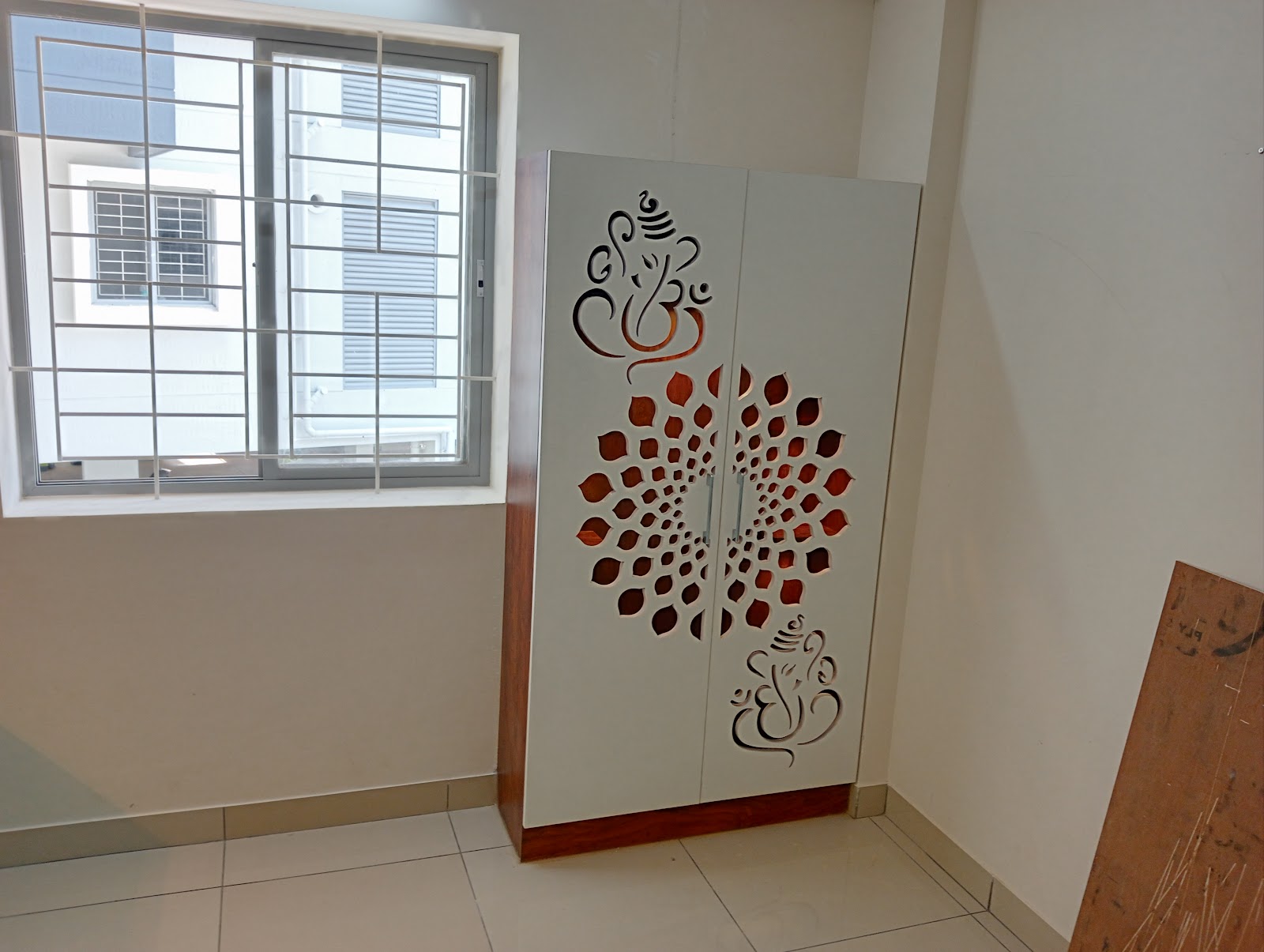 Standalone white pooja cabinet with laser-cut Ganesha motif and mandala jaali door panels on a wood-tone plinth base, set in an otherwise bare room.