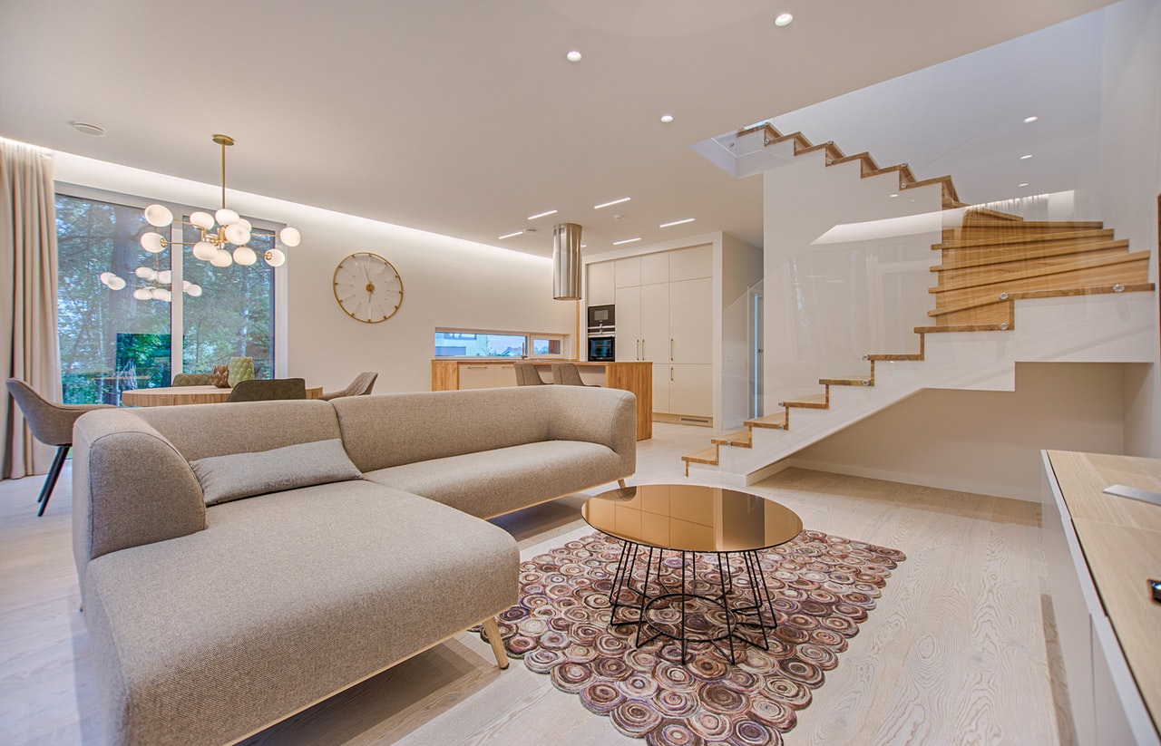 An open-plan living-dining-kitchen with a floating wood staircase, a beige sectional sofa on a circular patterned rug, a glass-top brass coffee table