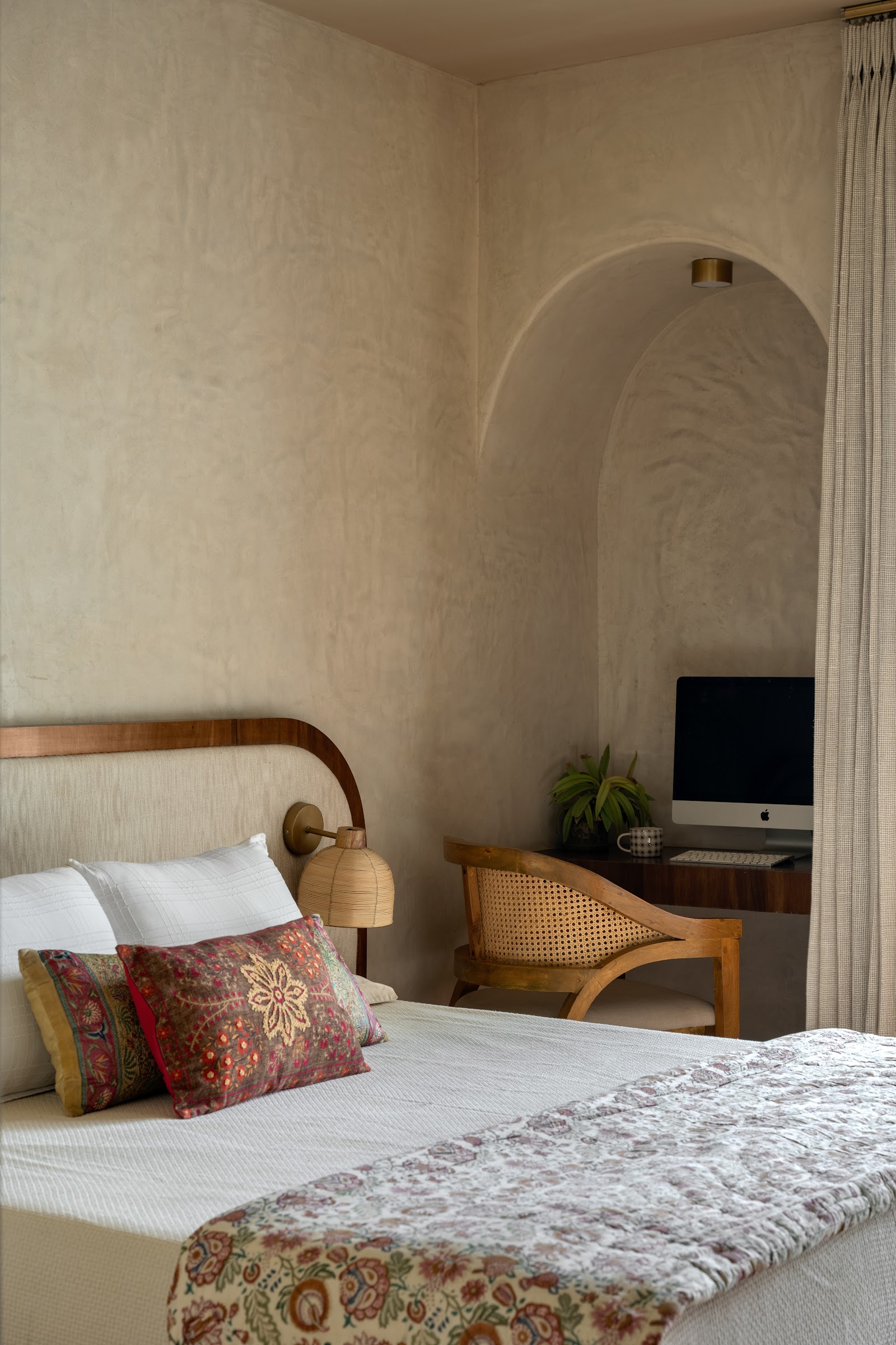 Bedroom with a curved wood-and-linen headboard and block-print cushions, opening to an arched lime-plaster niche with a built-in study desk and cane chair.