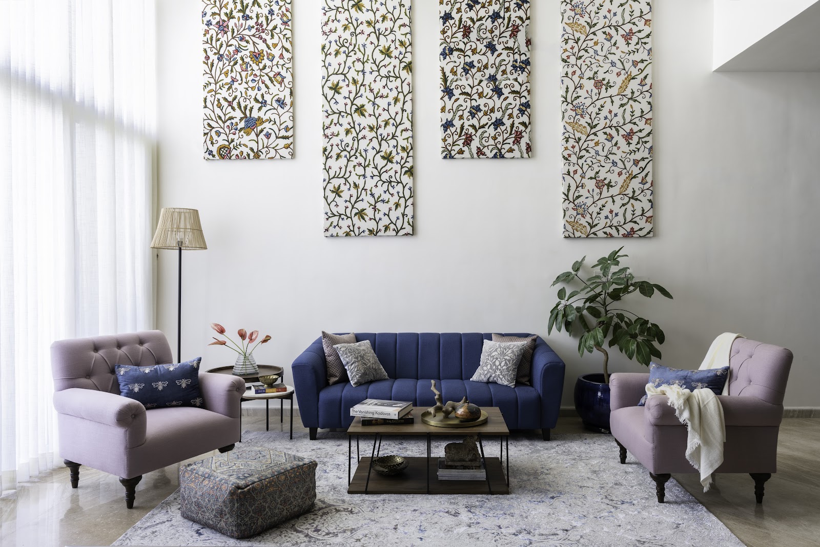 Double-height living room with a channel-tufted navy sofa, lilac tufted armchairs, a gallery of four floral crewel-embroidered wall panels, and a patterned rug.