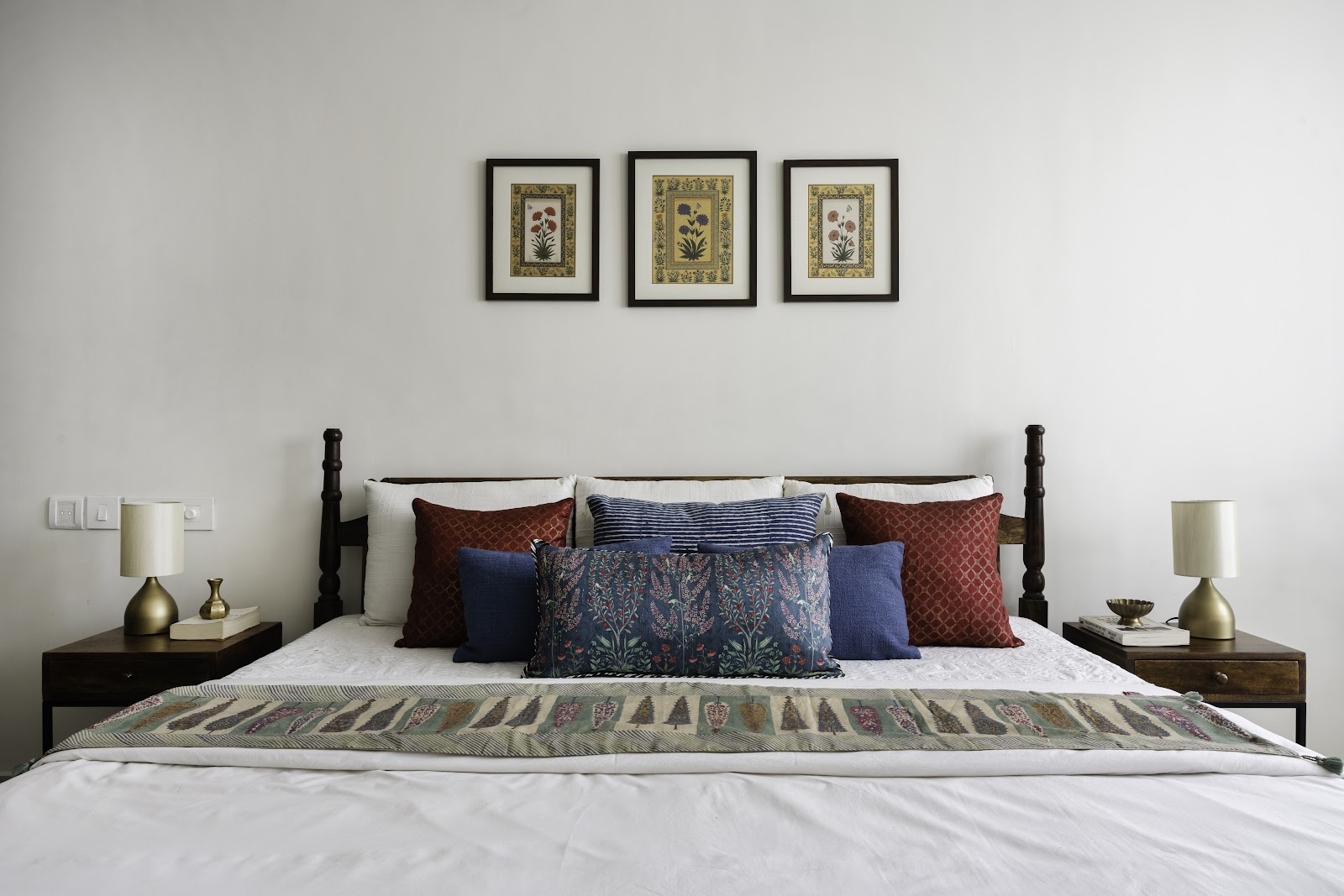 Bedroom with a dark wood four-poster bed dressed in red, blue and indigo block-print cushions, a folded embroidered throw at the foot, brass table lamps