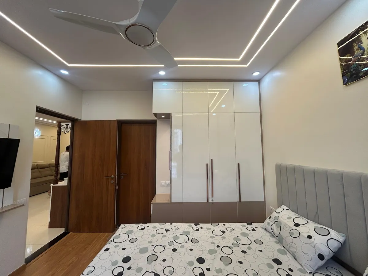 Bedroom with a grey fluted upholstered headboard beside a floor-to-loft high-gloss ivory wardrobe, under a false ceiling cut with continuous LED profile lines
