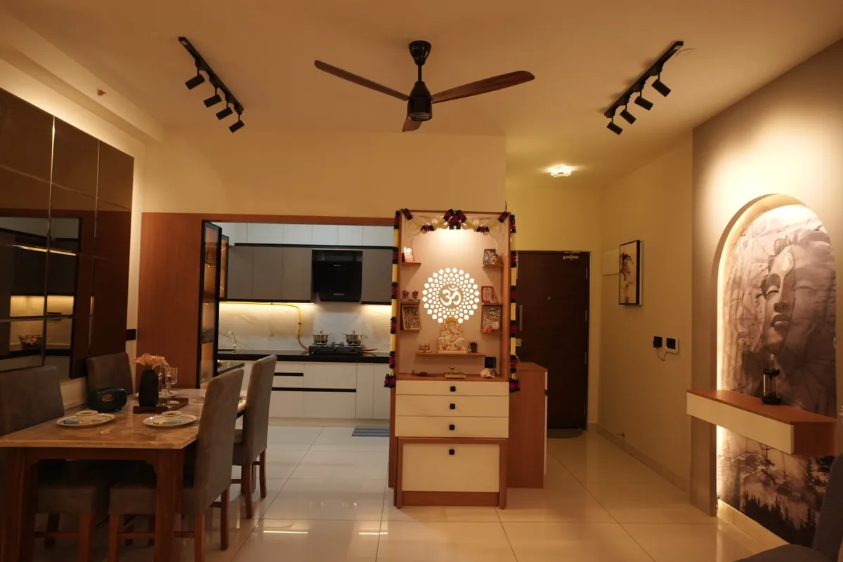 Warm evening view across an open plan where a freestanding pooja unit with a backlit Om cut-out and marigold torans splits the dining table from the kitchen