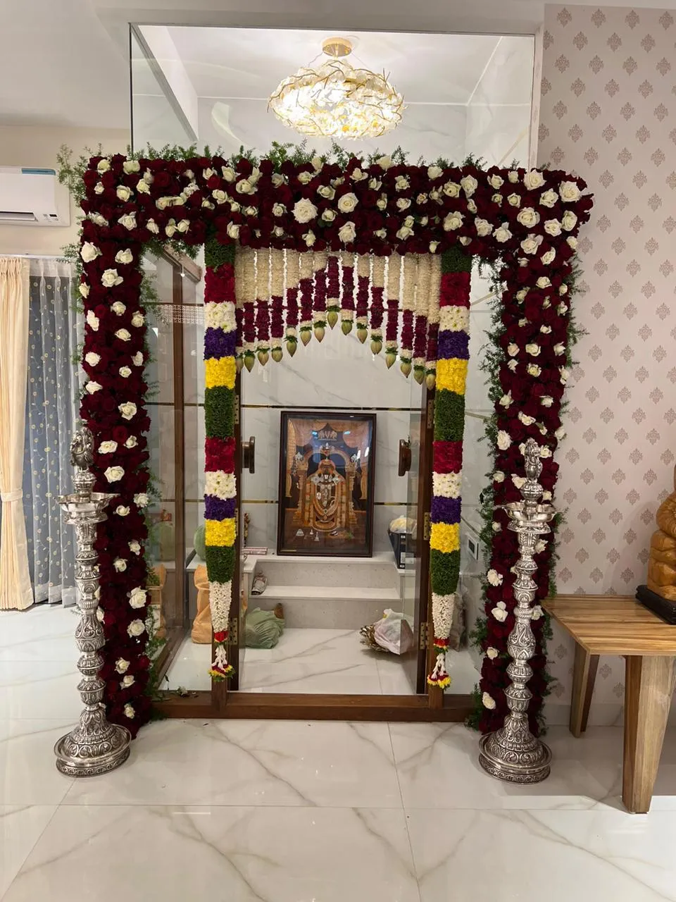 Glass-fronted home pooja niche dressed for a function — rose-and-marigold garlands framing the teak-and-glass doors