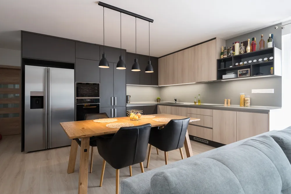 L-shaped charcoal-and-wood kitchen with a wooden dining table under black pendant lights, opening onto a grey sofa in the foreground.
