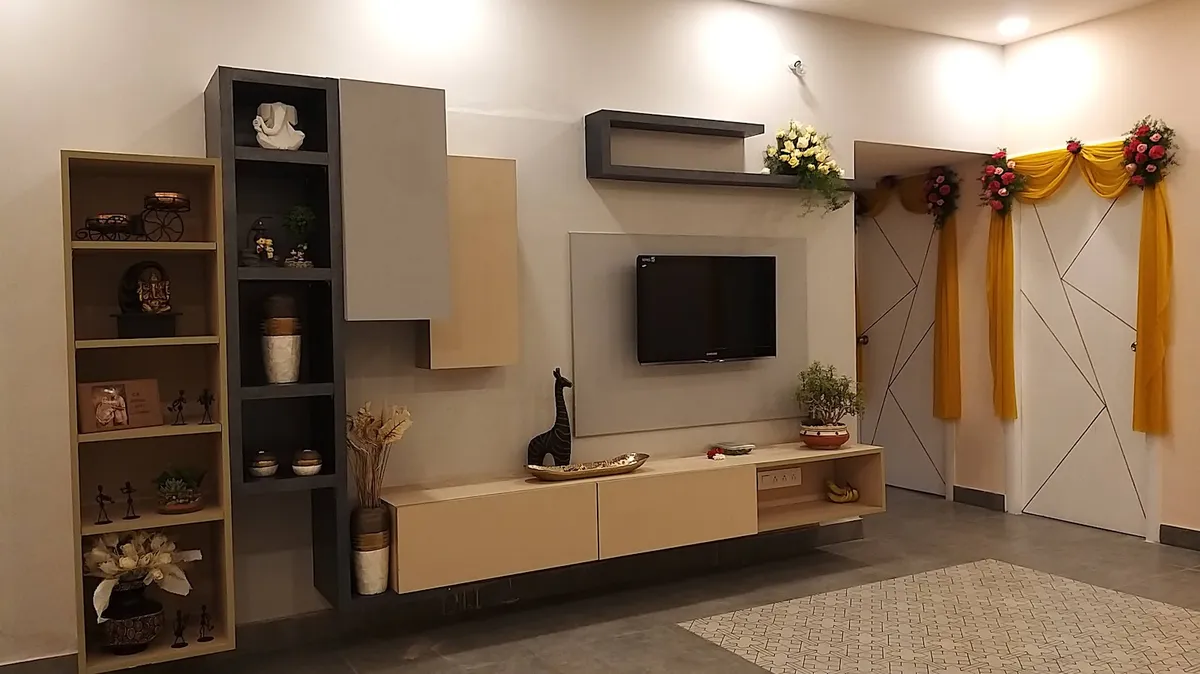 Living room TV wall on a housewarming day: beige-and-charcoal laminate console with a floating shelf and open display boxes holding brass curios and Ganesha