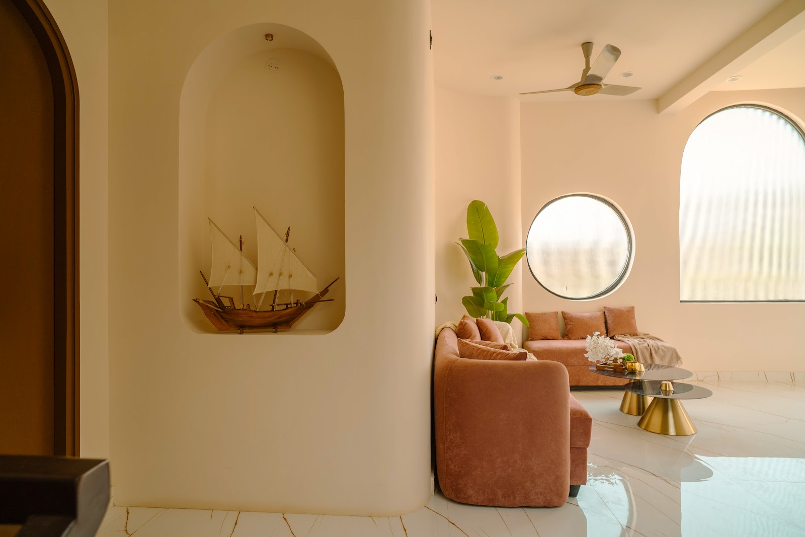 Living room with curved plastered walls, an arched wall niche displaying a wooden ship model, dusty-rose velvet sofas, brass conical nesting coffee tables