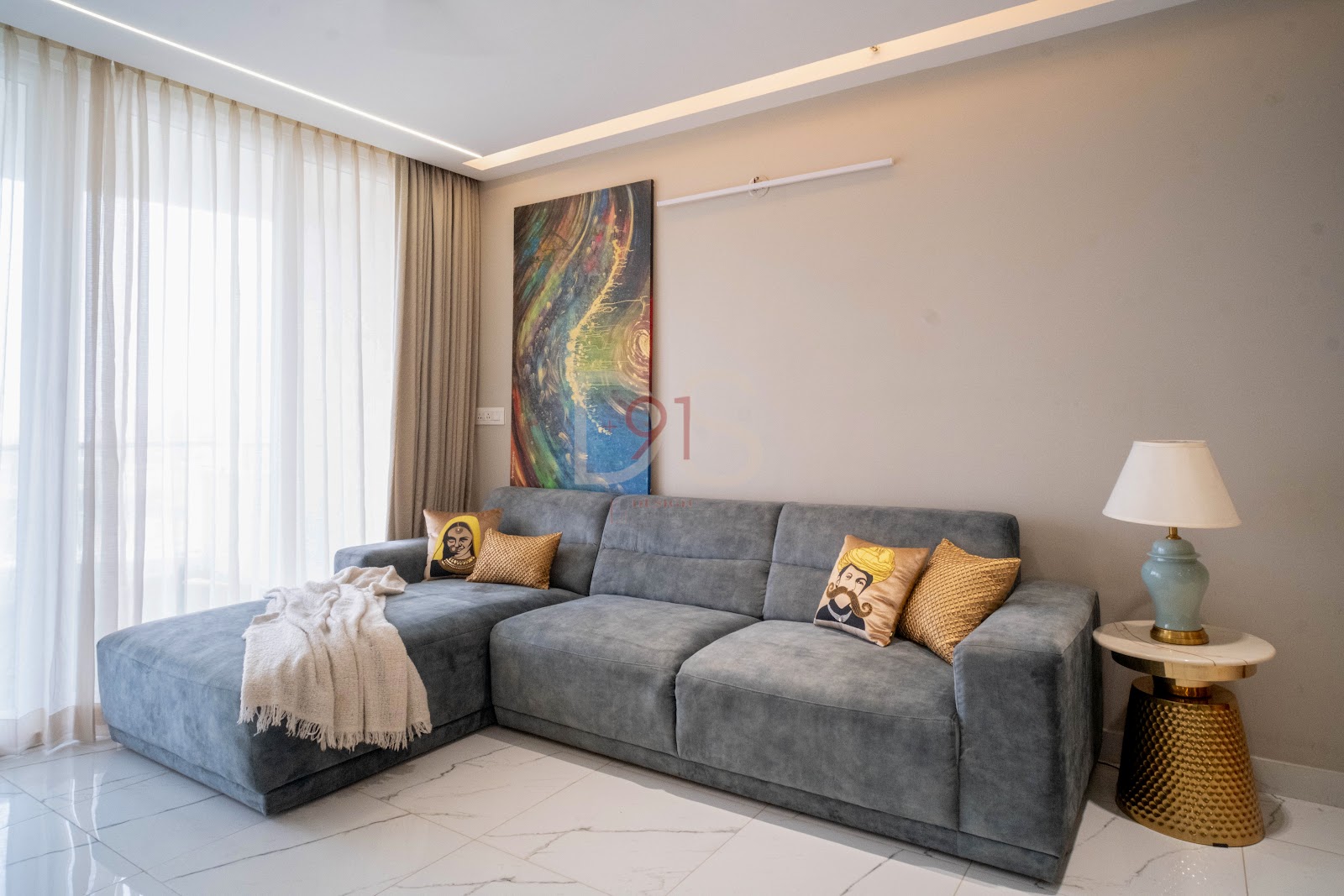 Grey L-shaped sectional sofa in a living room with a cove-lit ceiling, marble-look tile flooring, and gold turban-motif cushions against a beige accent wall.