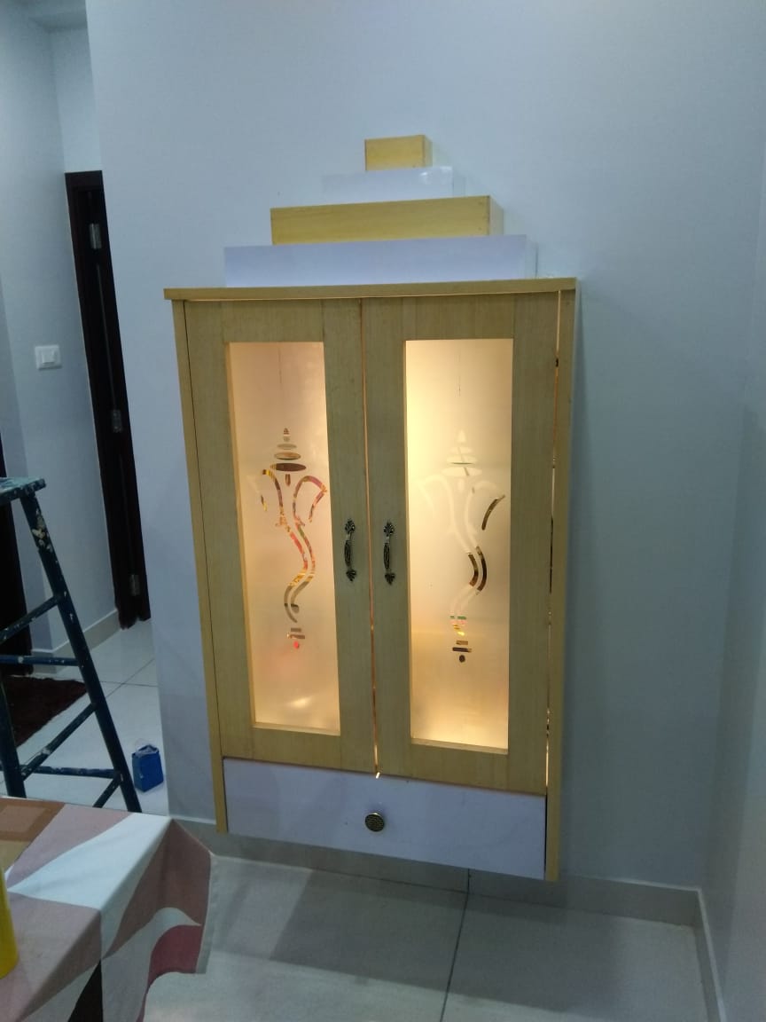 Wall-mounted pooja cabinet with backlit frosted glass doors etched with a Ganesha motif, a tiered wood pelmet top, and a lower drawer with a brass knob.