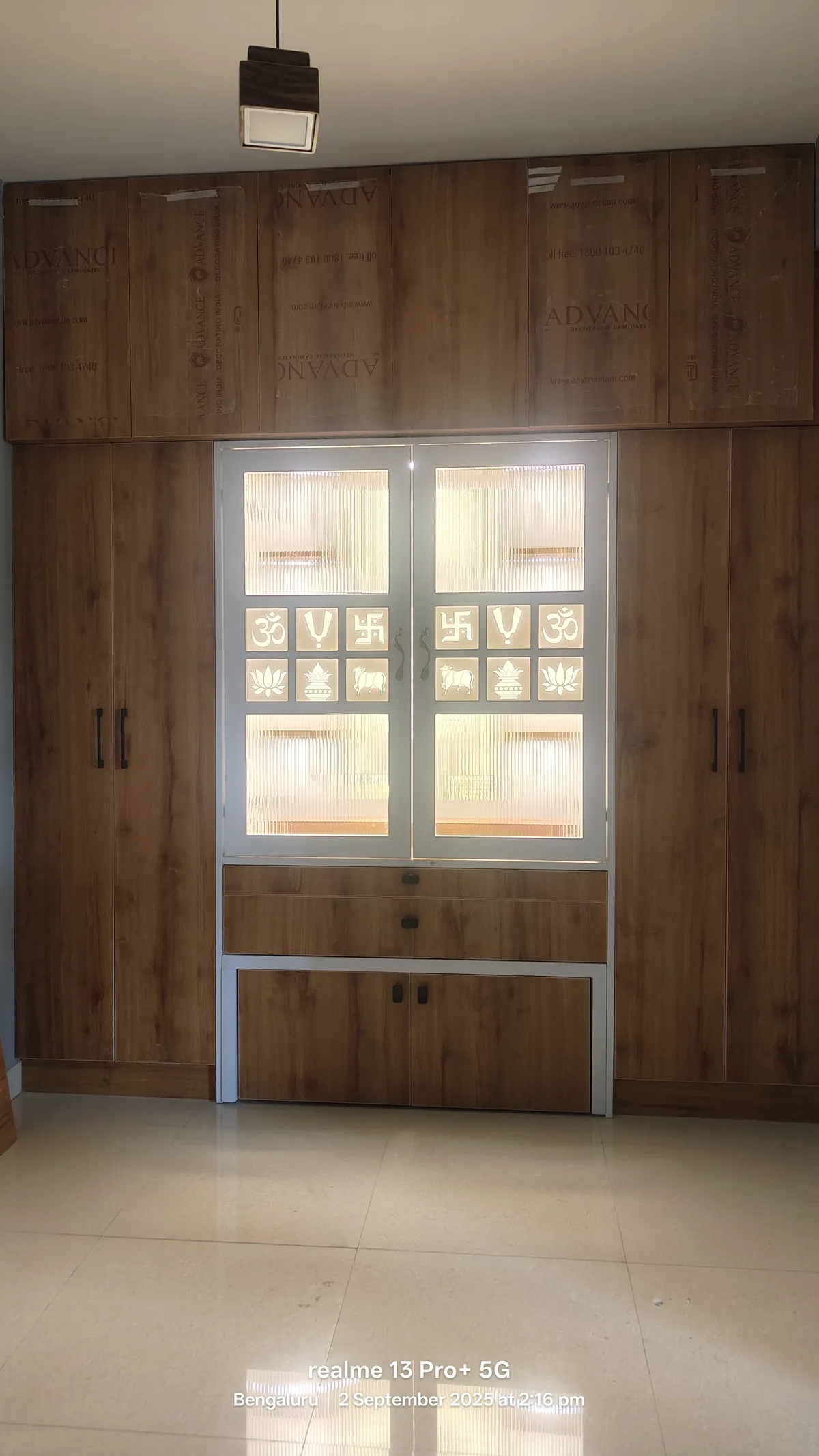 A built-in wooden pooja unit with backlit fluted-glass doors featuring Om, swastik and lotus motifs, flanked by full-height wardrobe cabinets