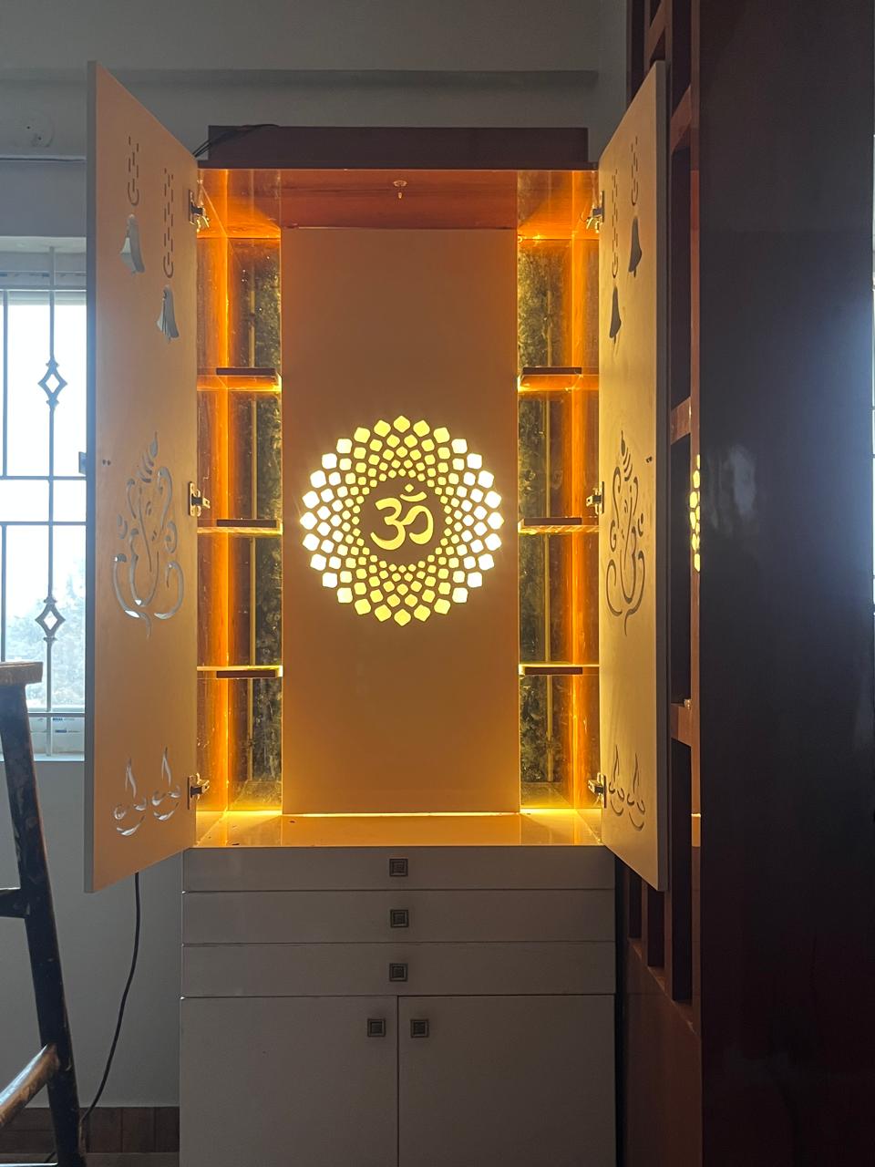 Backlit pooja unit with a laser-cut golden Om mandala centrepiece and Ganesha cut-outs on the cabinet doors, set above a white storage base