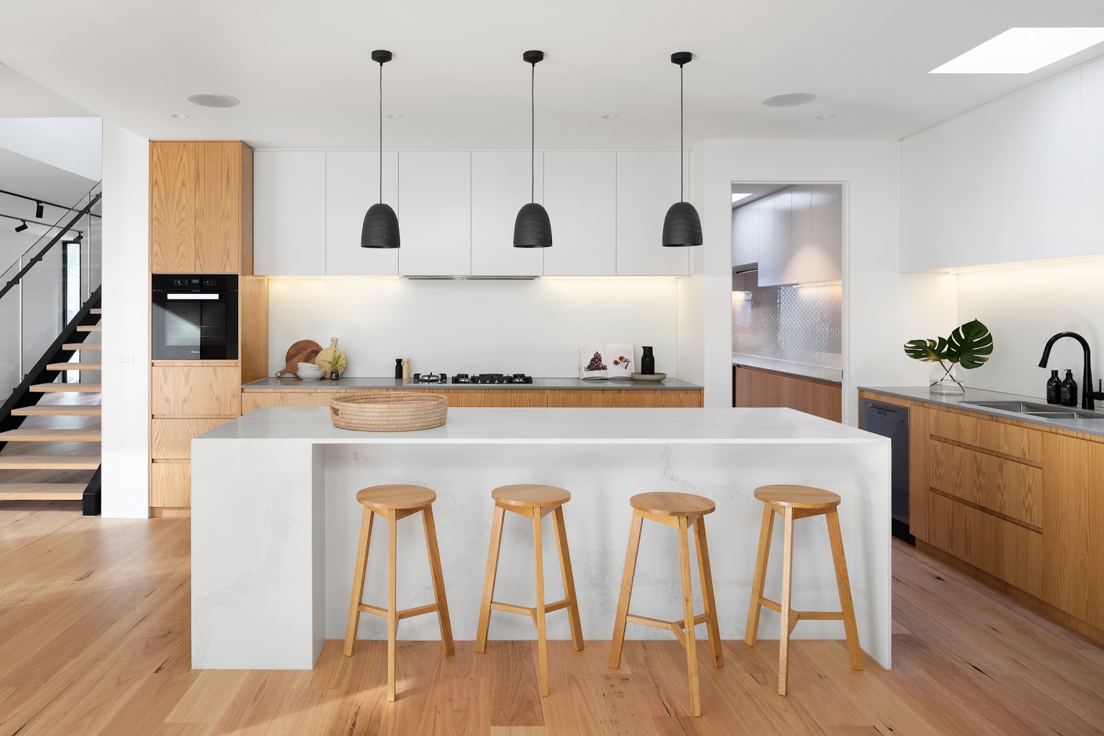 Kitchen with a white waterfall-edge island, four oak counter stools, and three black dome pendant lights over an oak-cabinet run with integrated oven.