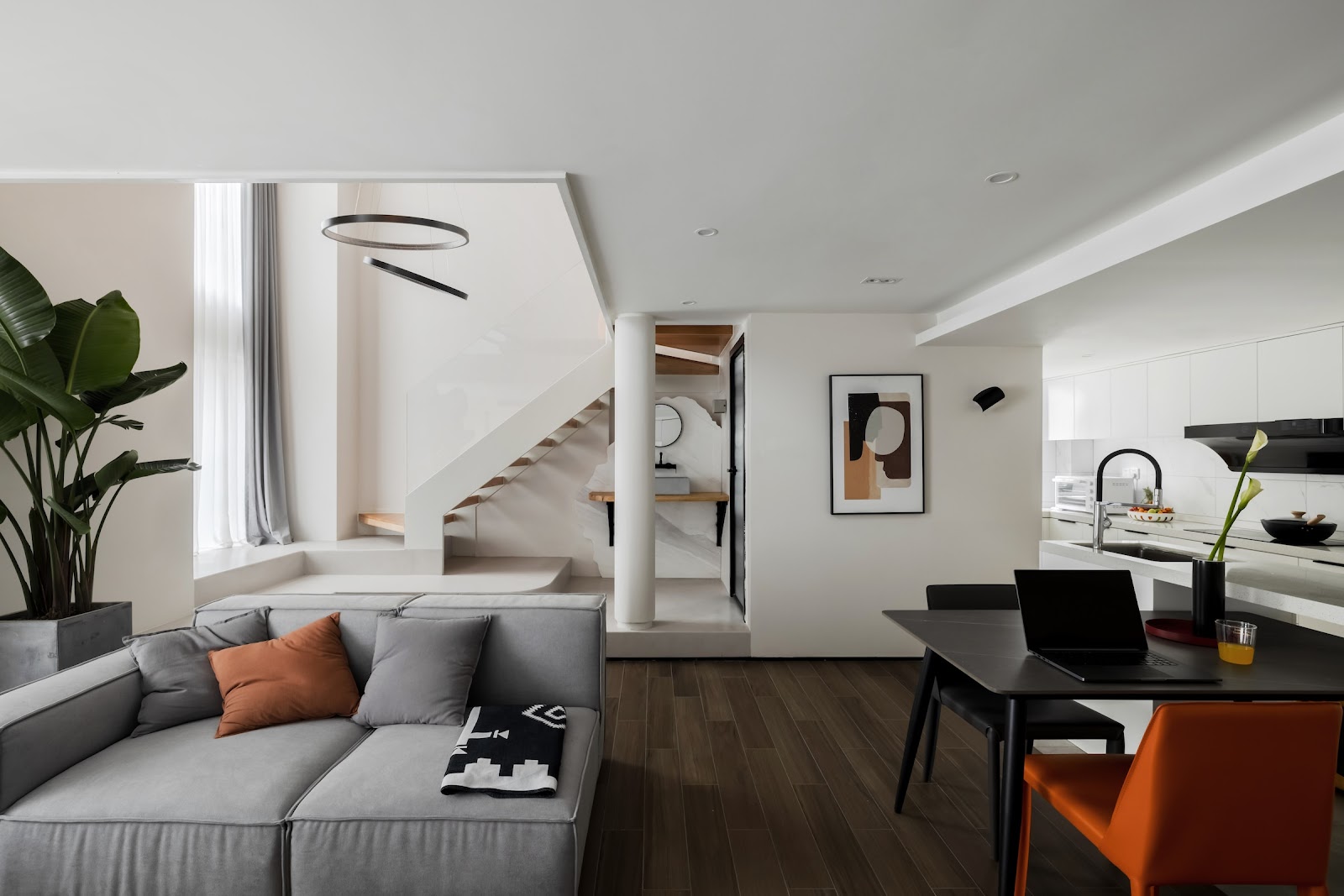 Open-plan living-dining-kitchen loft with a floating staircase, grey modular sofa with an orange throw pillow, and an orange dining chair accent.