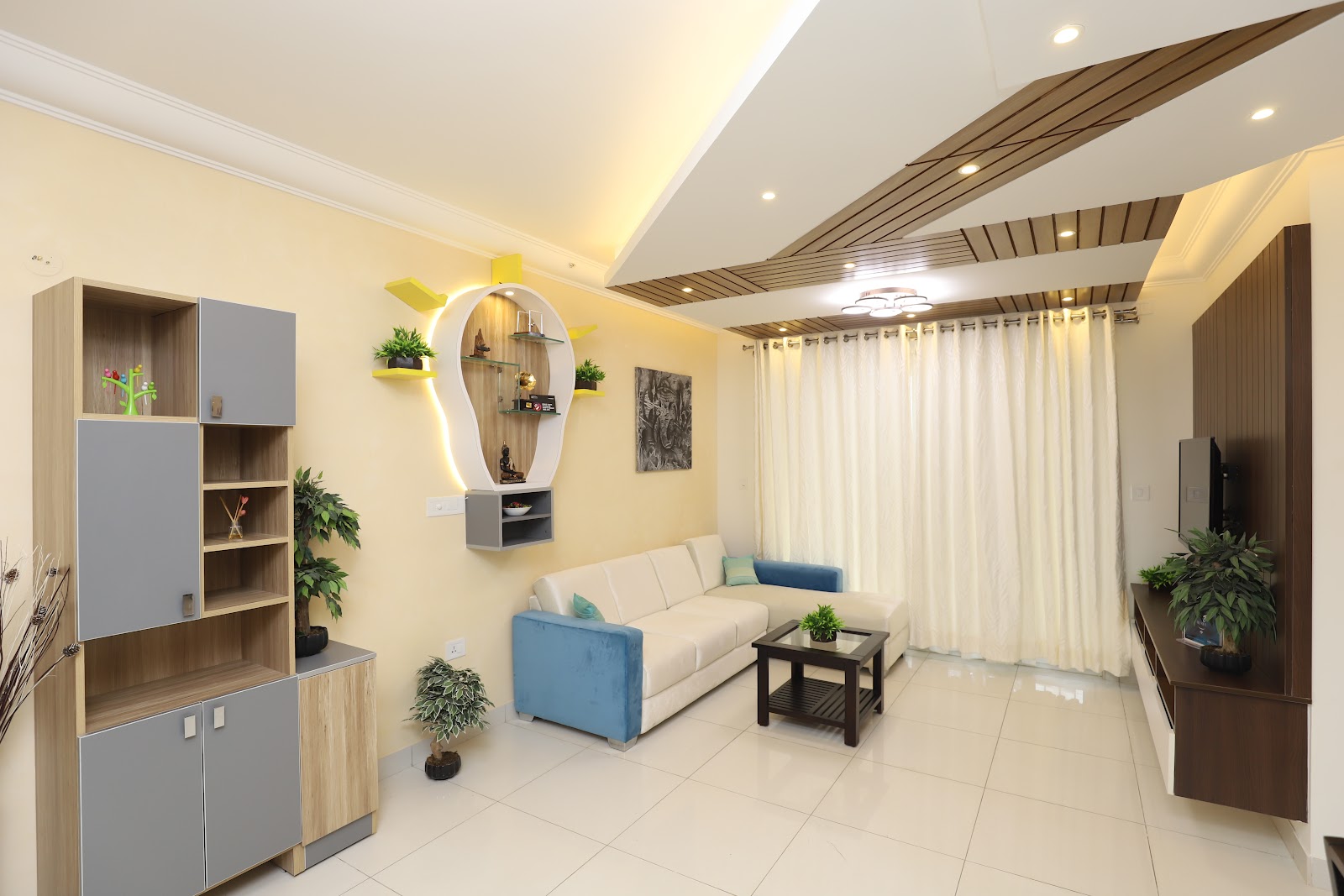 Living room with a wood-slat angled false ceiling, a cream-and-blue L-shaped sofa and a wall-mounted yellow-accented pooja niche with a Buddha statue.