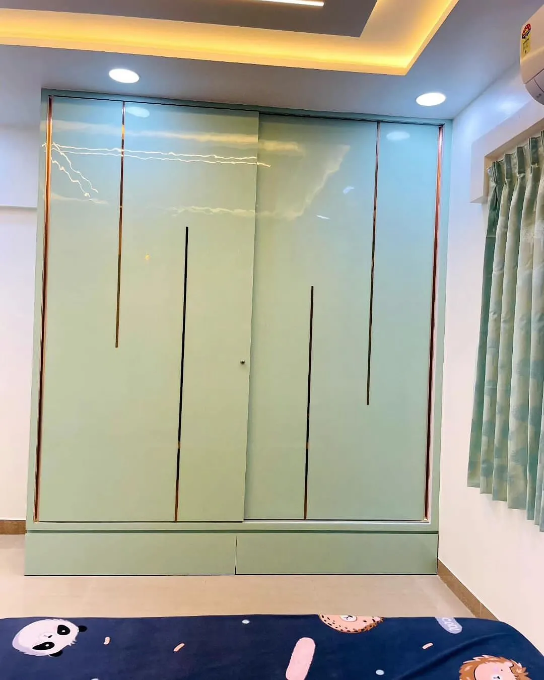 Mint-green high-gloss sliding wardrobe with rose-gold profile grooves and two base drawers, shot across a bed made up in panda-and-lion print bedding