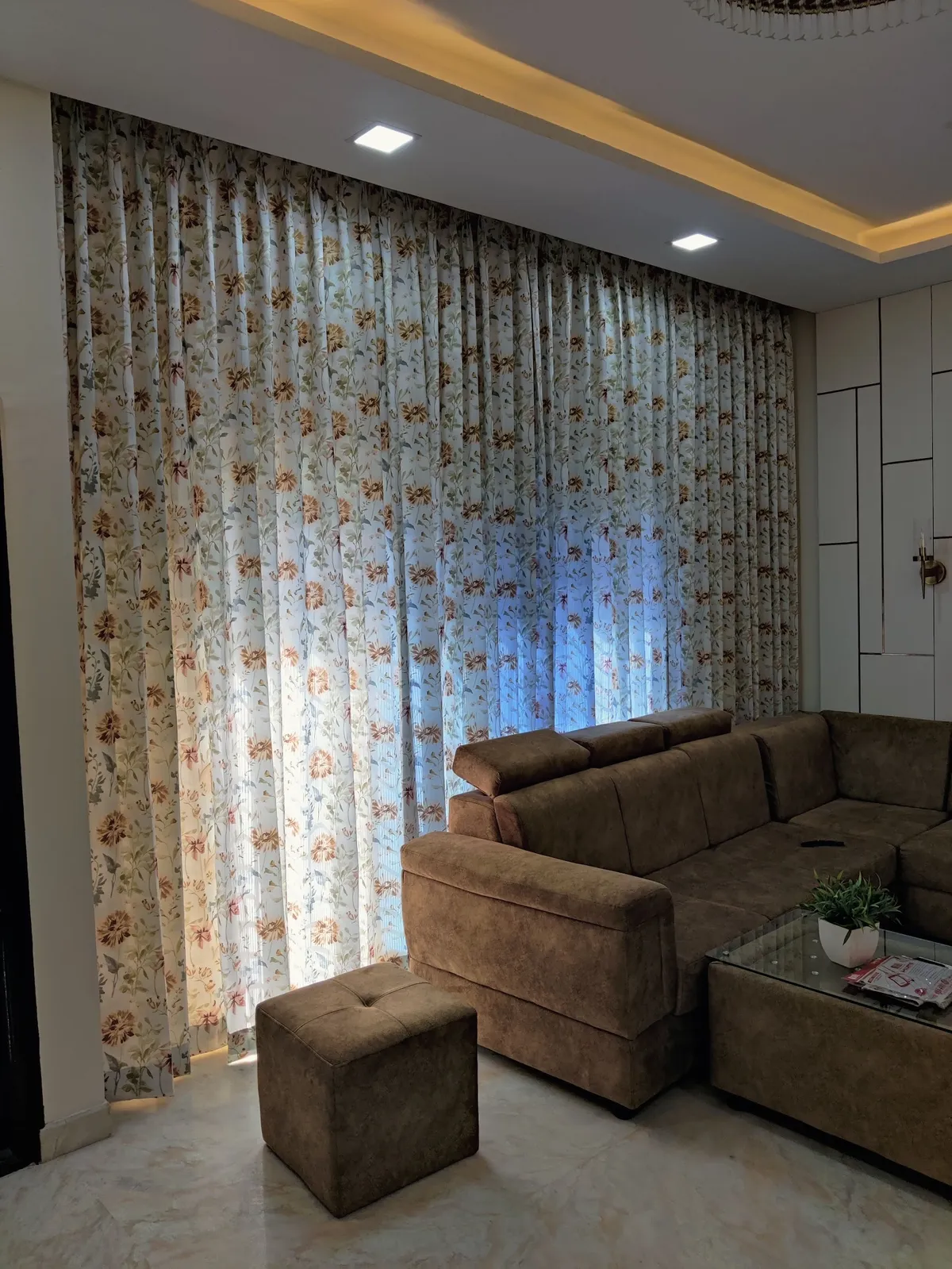 Living room with floral-print sheers drawn across an entire wall, a brown suede L-sofa with matching cube ottoman and a glass-top storage coffee table under a