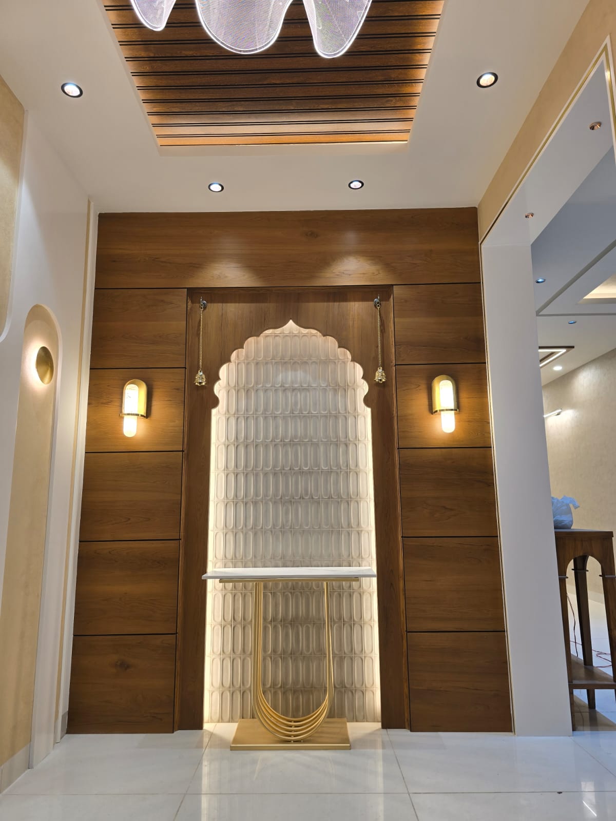 Pooja alcove with an arched, backlit textured tile niche framed by walnut wood panelling, a brass console table, and hanging temple bells.