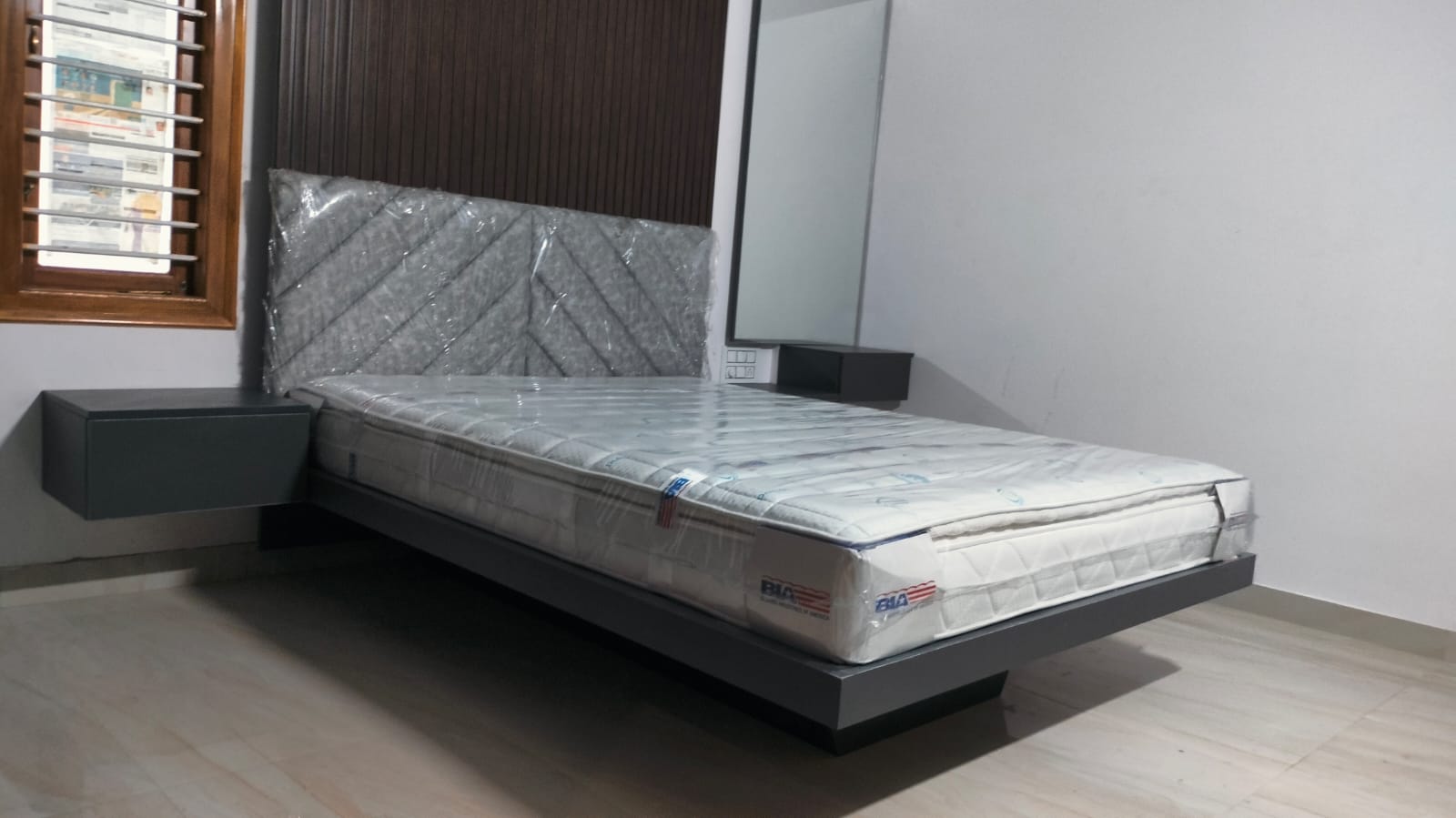 Bedroom with a floating platform bed against a dark wood-slat headboard wall and matching wall-mounted nightstands; mattress still in delivery wrap.